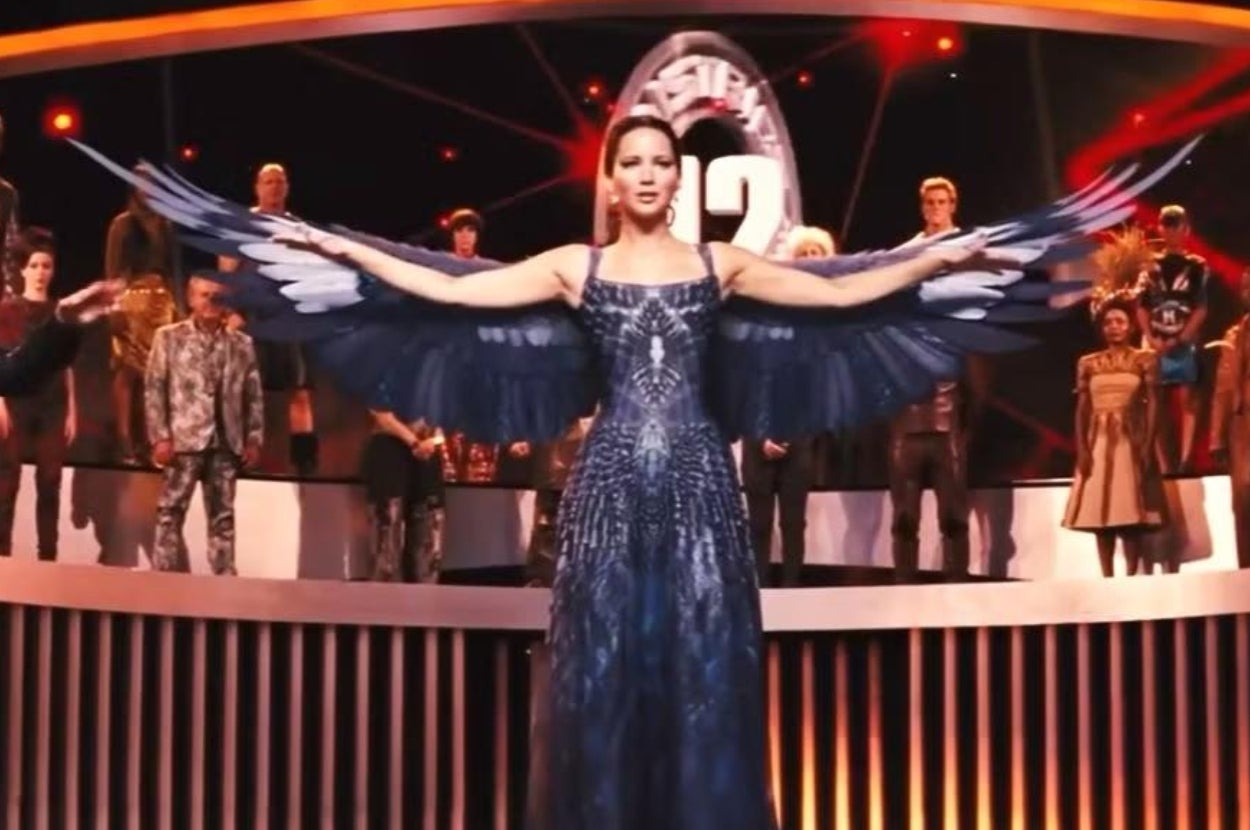 Actor in a detailed gown with wing extension on stage, arms outstretched, portraying a character in a dramatic event