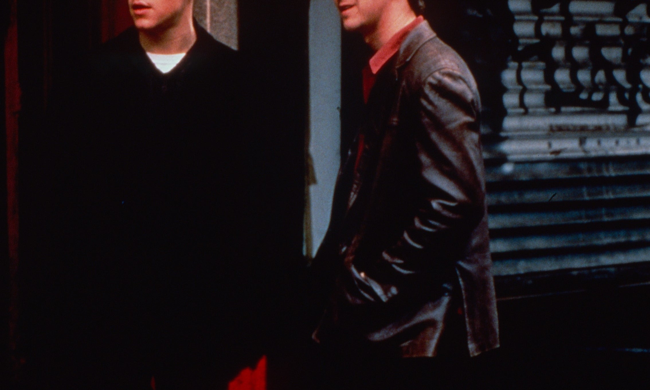 Two people stand outside, casually dressed, one in a coat and the other in a leather jacket, looking off into the distance