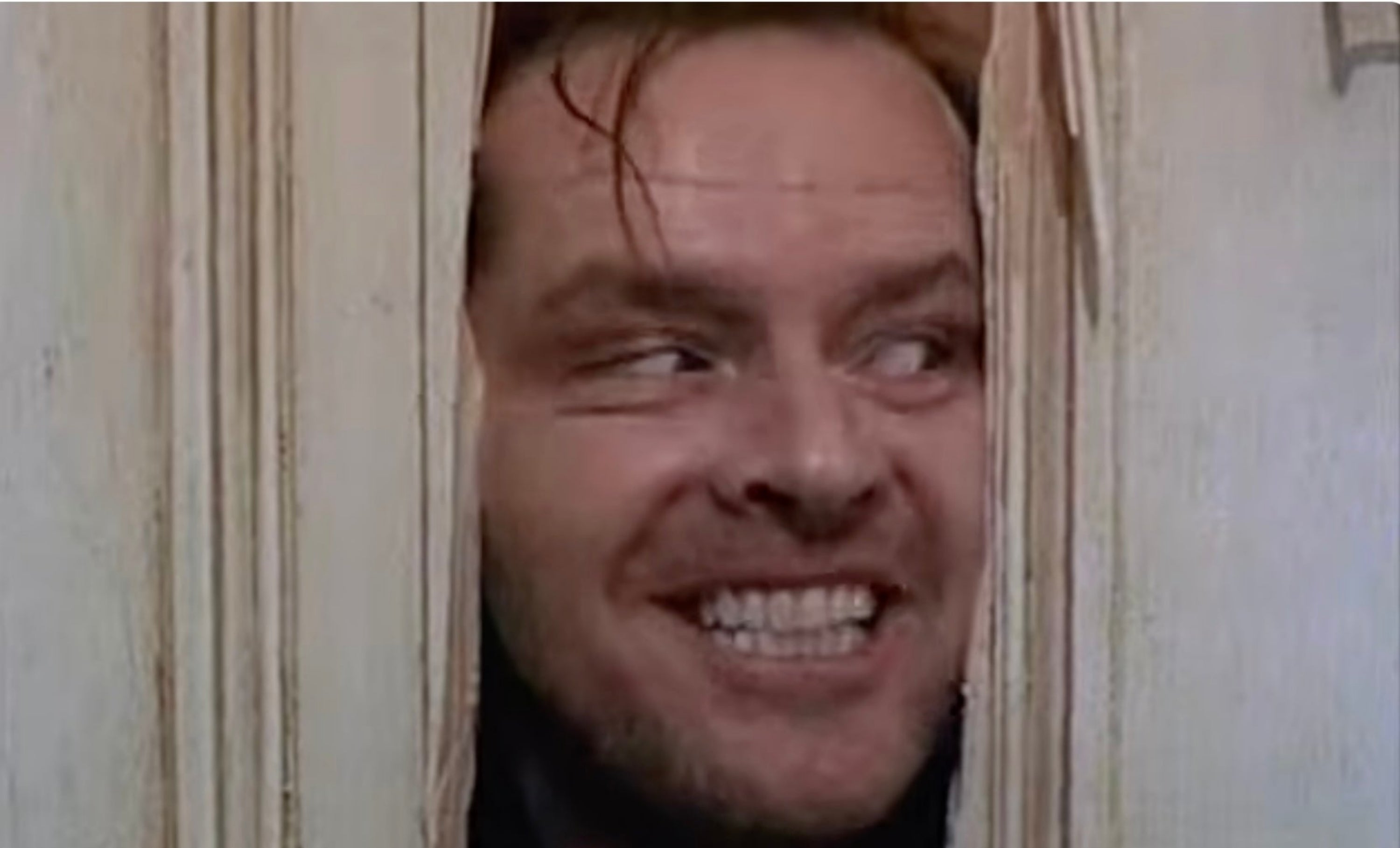 Person looking through a broken door with a wide grin, head slightly tilted. Scene from a famous movie
