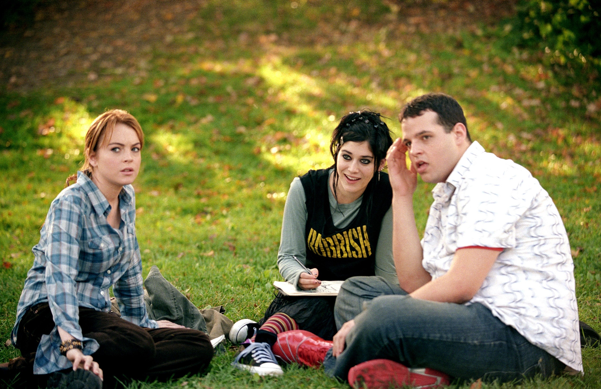 Three people sitting on grass in a park; one writes in a notebook. Casual, relaxed scene