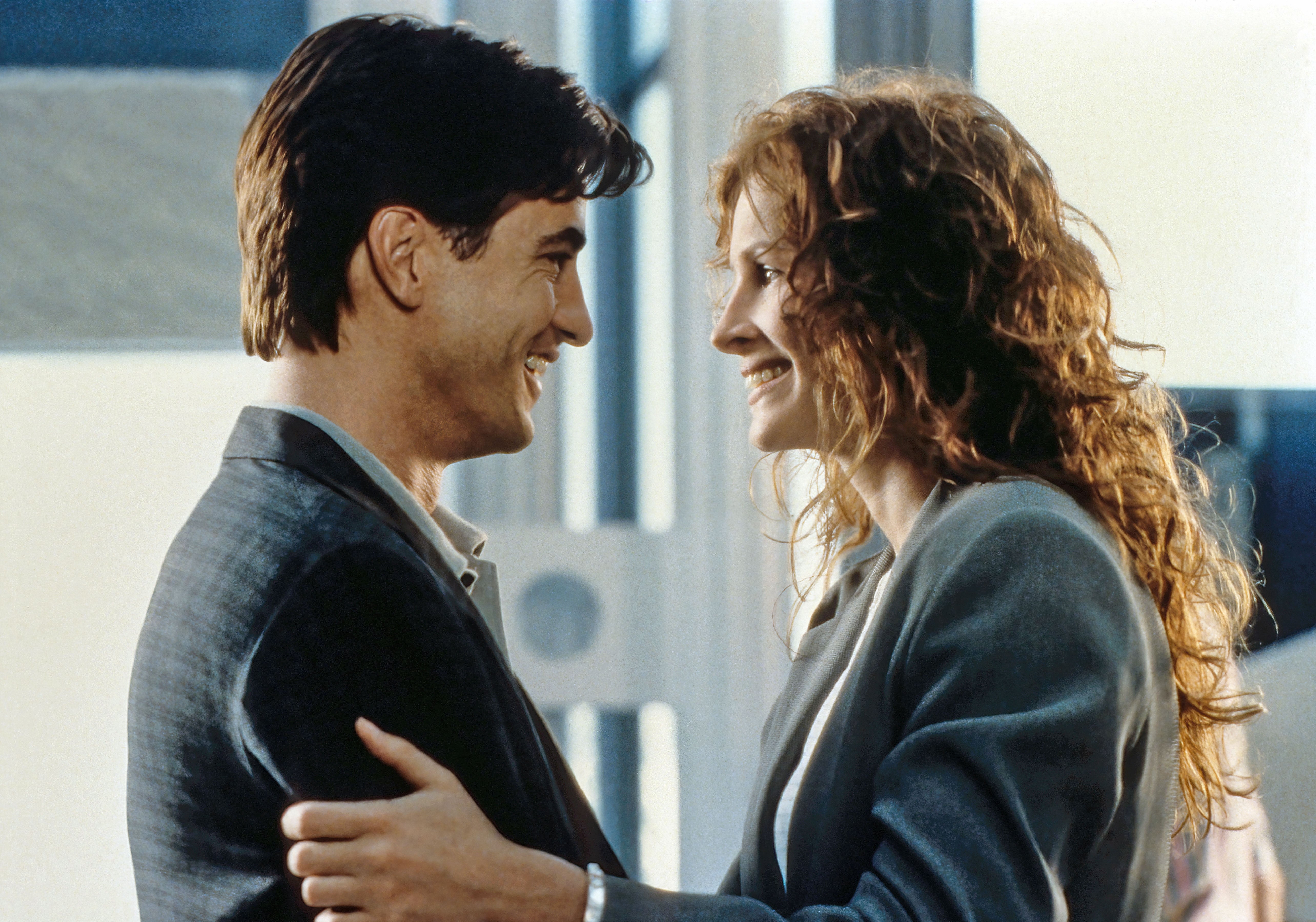 A man and woman smile at each other closely, both wearing business attire. They seem to be in an office setting