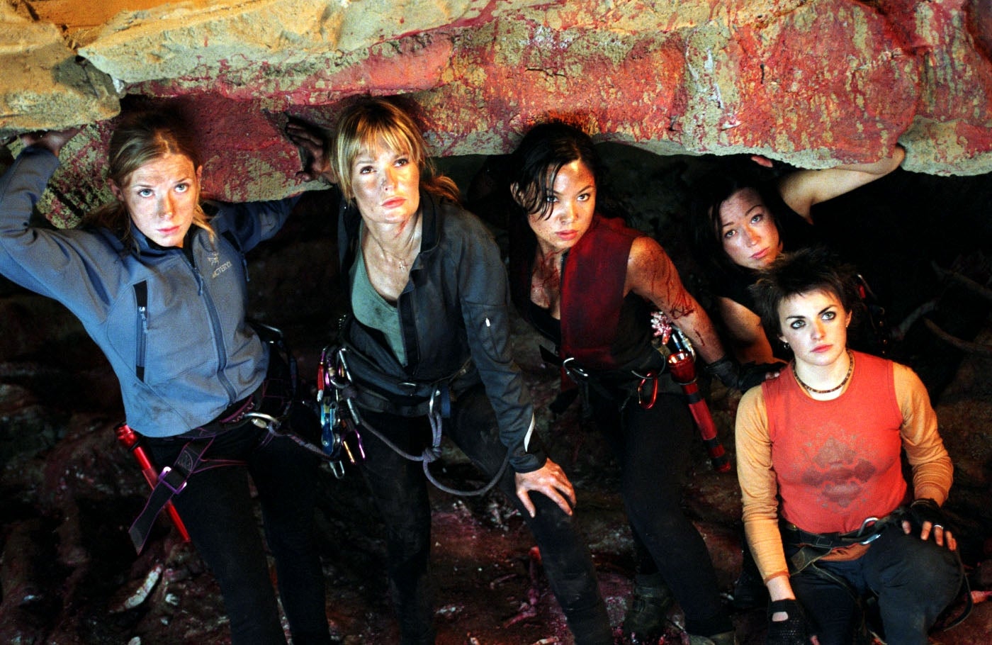 Five women in adventure gear pose in a rugged cave setting, each showing determined expressions and readiness for exploration