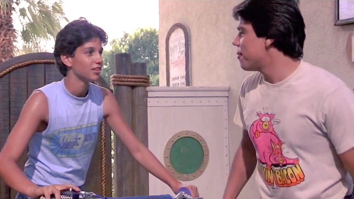 Two teenagers stand facing each other, one with short hair in a tank top, the other in a graphic T-shirt with a cartoon character