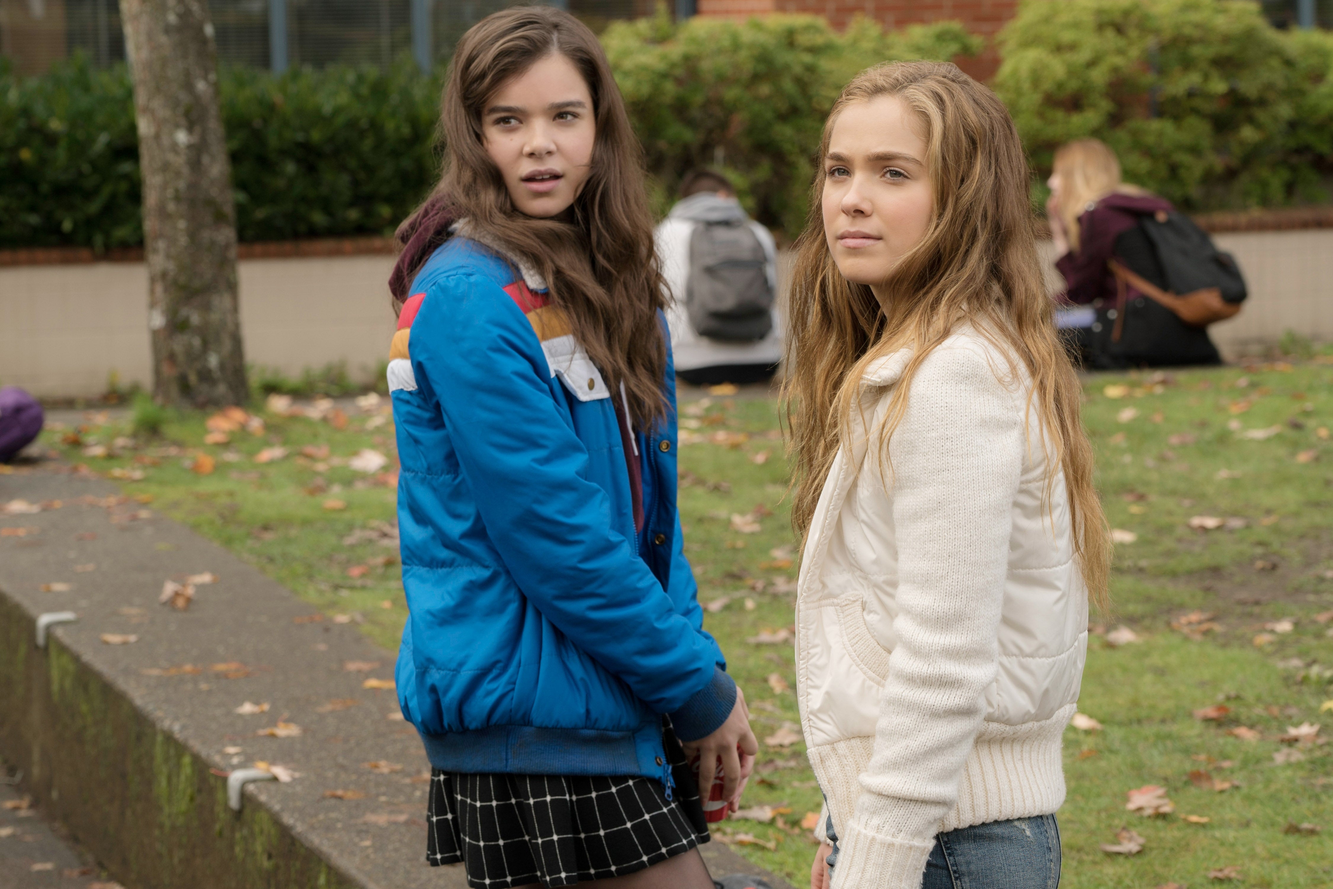 Two young women standing outdoors. One wears a blue jacket and plaid skirt, the other a white jacket. Background shows a park setting
