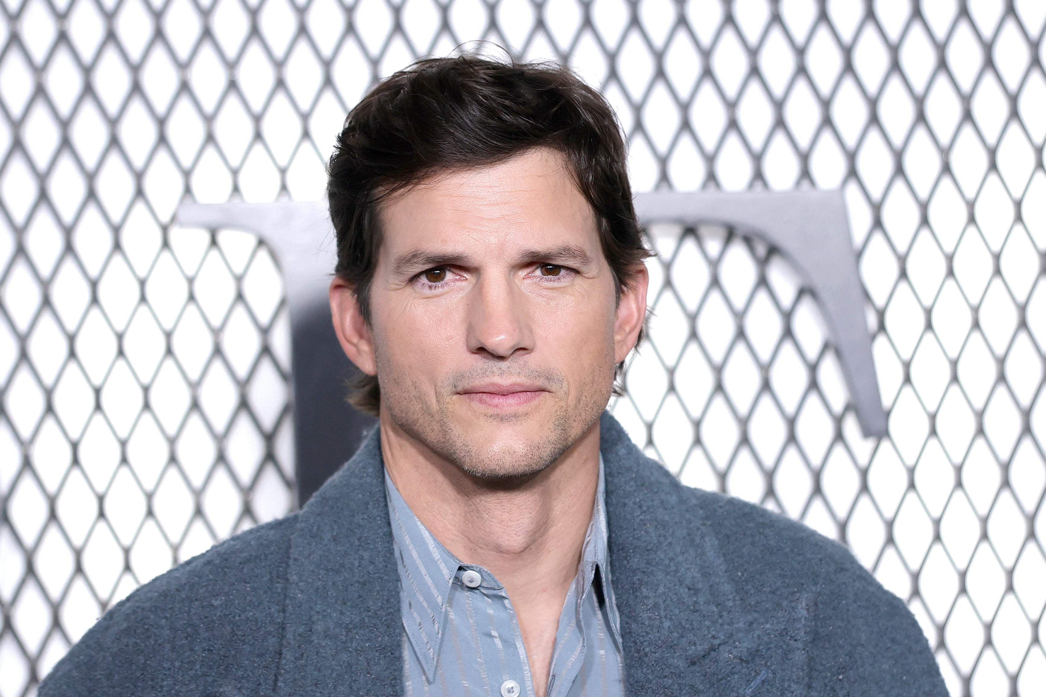 A person in a stylish, casual outfit stands in front of a net-patterned backdrop, at a media event