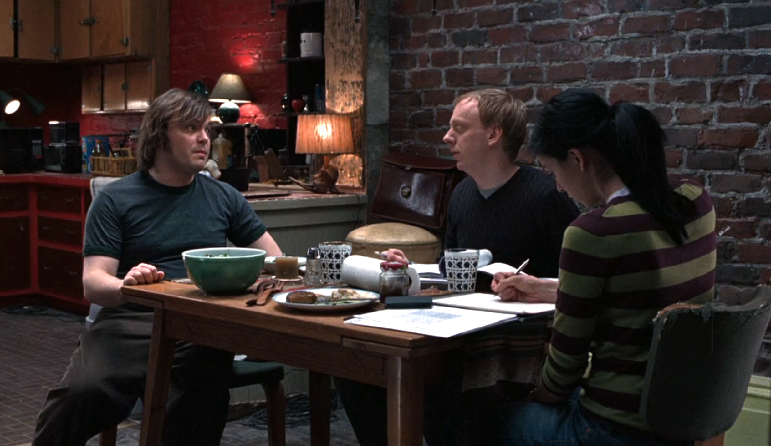 Three people sit at a wooden table with papers, cups, and food, engaged in conversation in a cozy room with brick walls and warm lighting