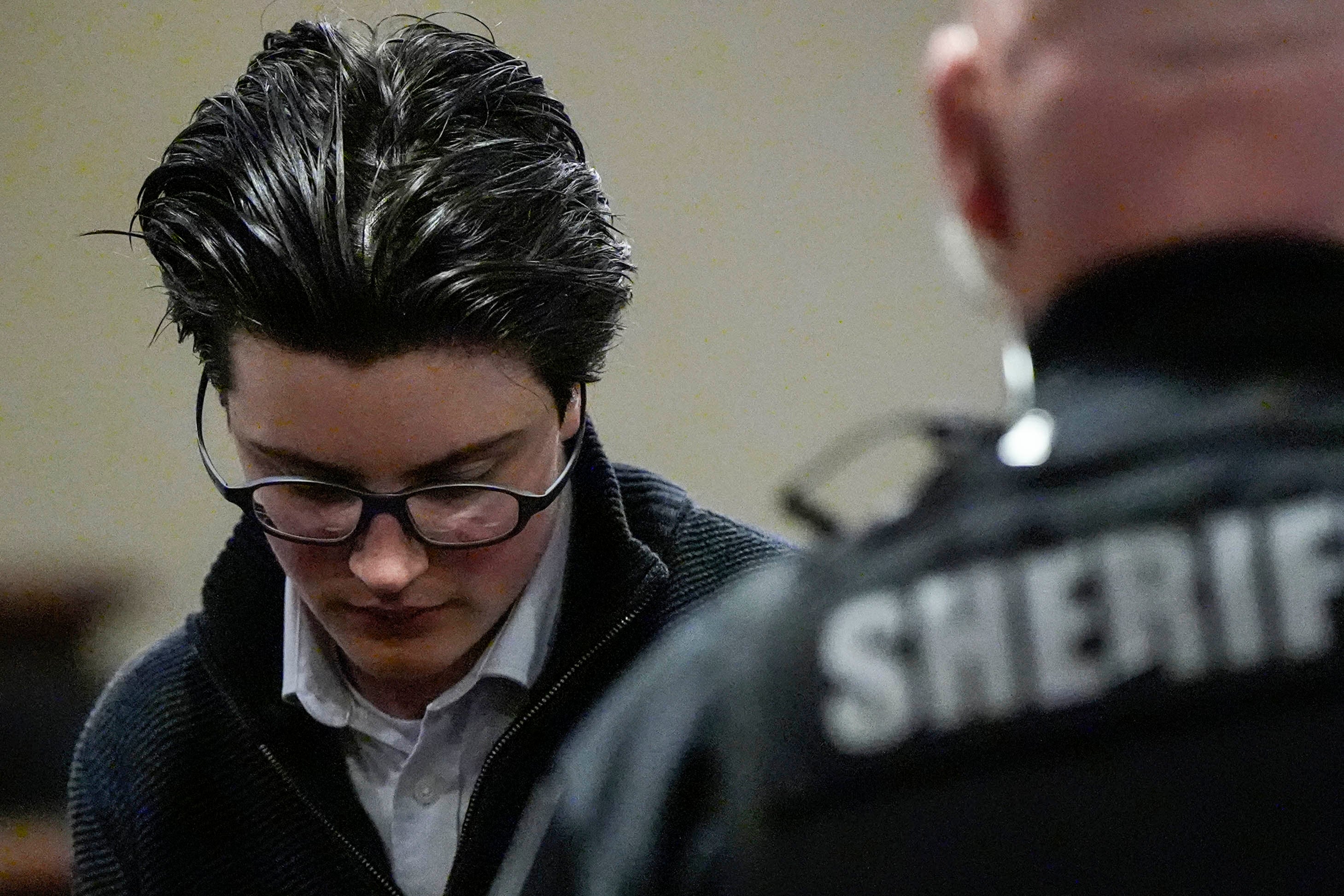 Person in glasses and a dark sweater looks down, next to a sheriff in uniform, in what appears to be a courtroom setting