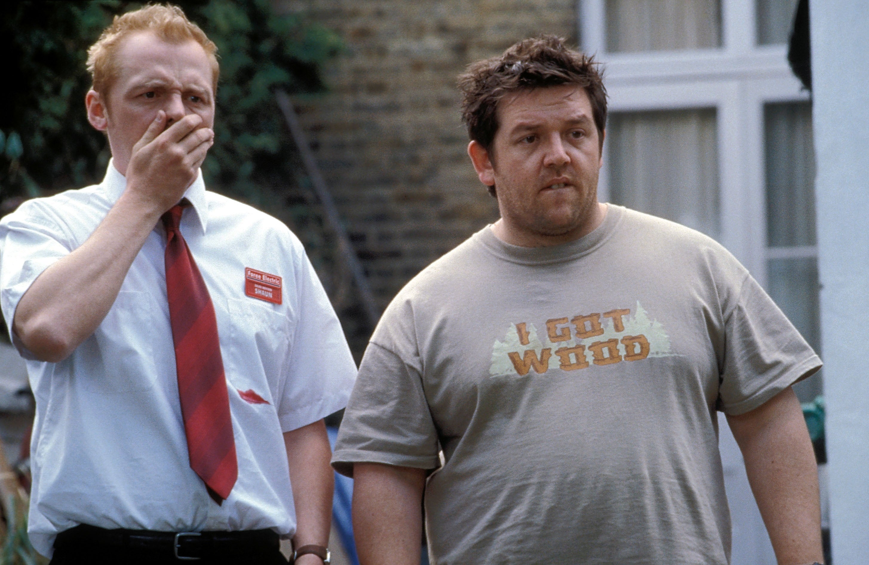 Two men stand outdoors. One wears a shirt with a humorous text, and the other appears surprised, with his hand over his mouth