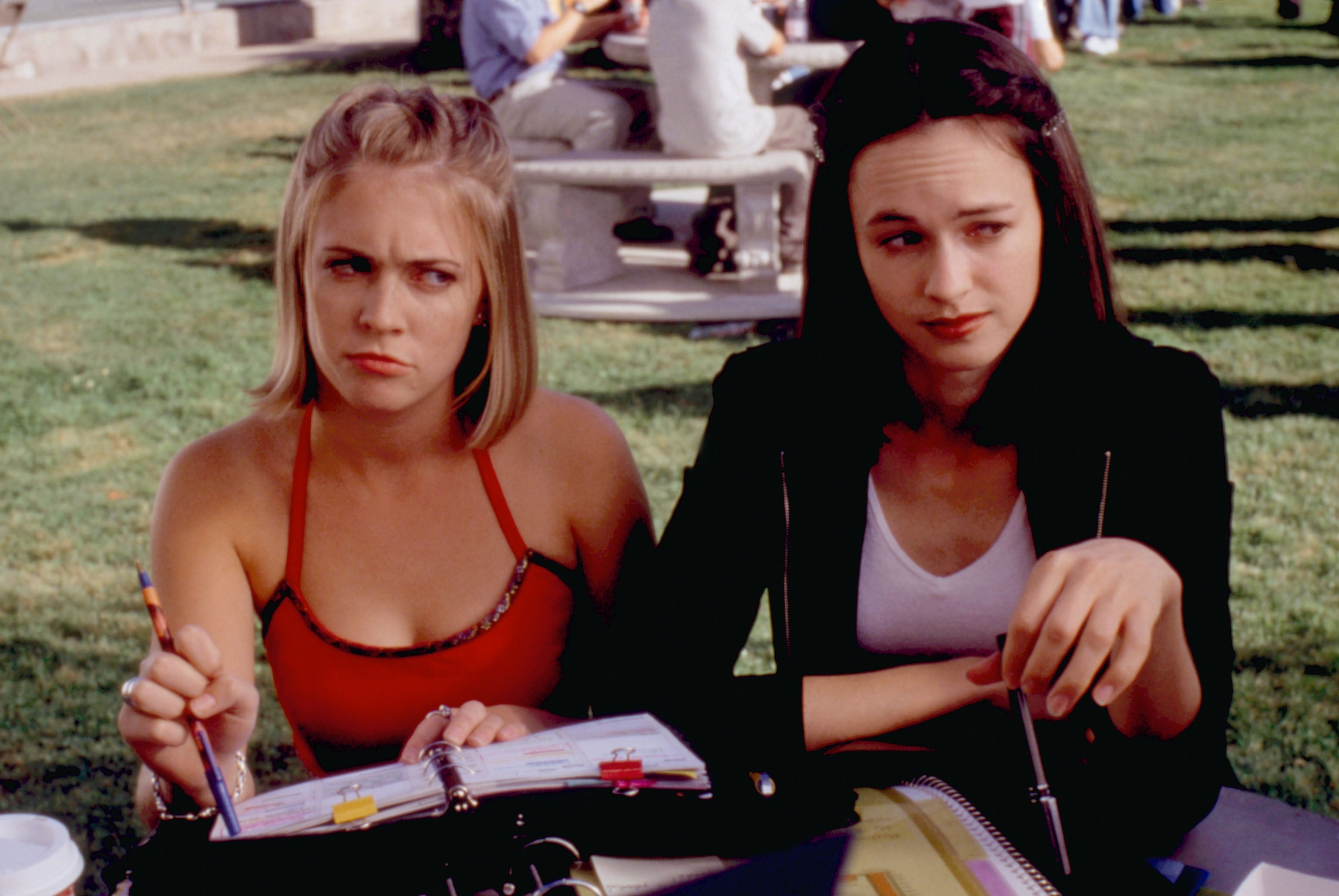 Two young women sit outdoors with open notebooks, appearing thoughtful and engaged, as if discussing or studying together