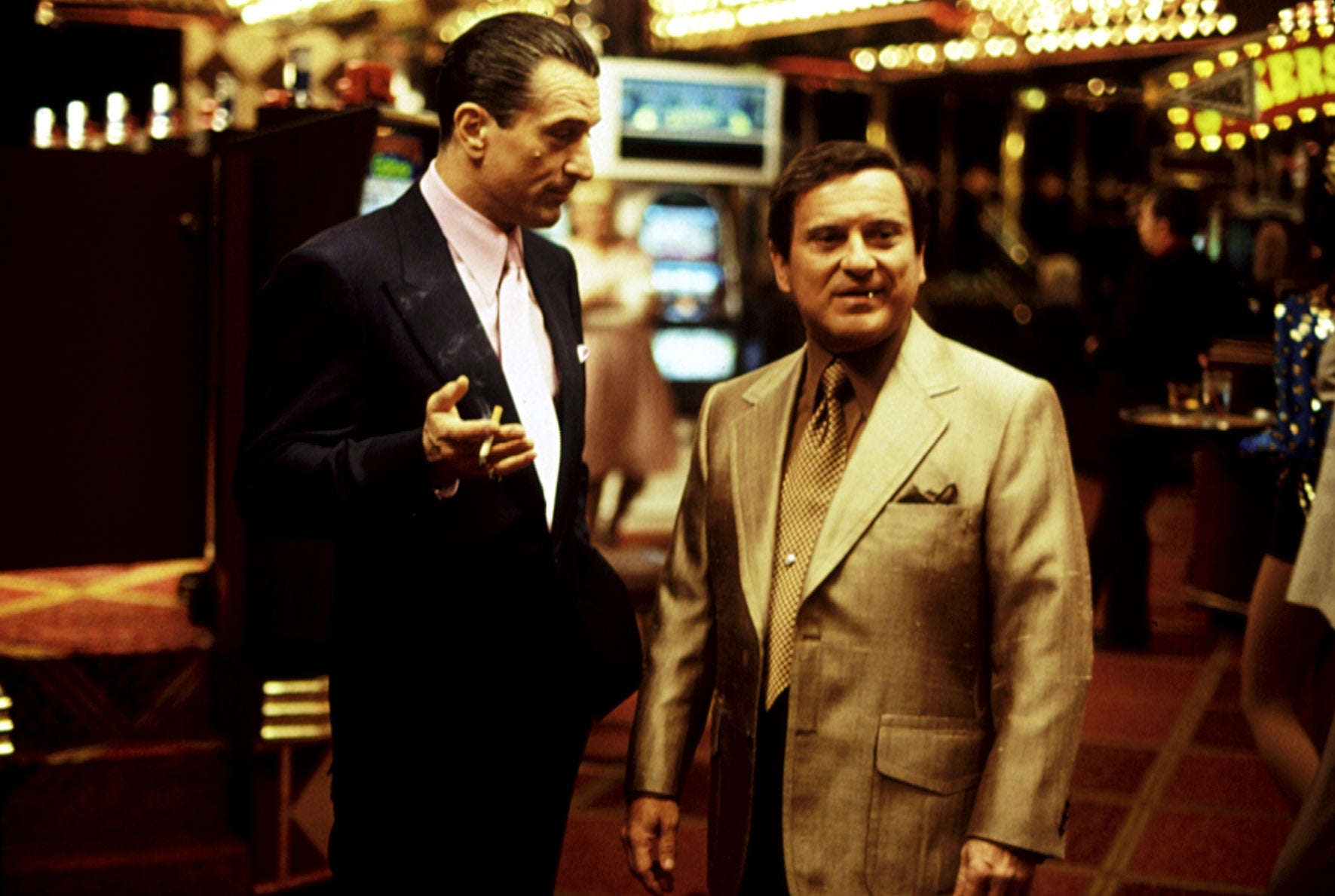 Two actors in a casino scene from a movie, wearing 1970s-style suits. One has a cigarette, while the other looks attentively at him