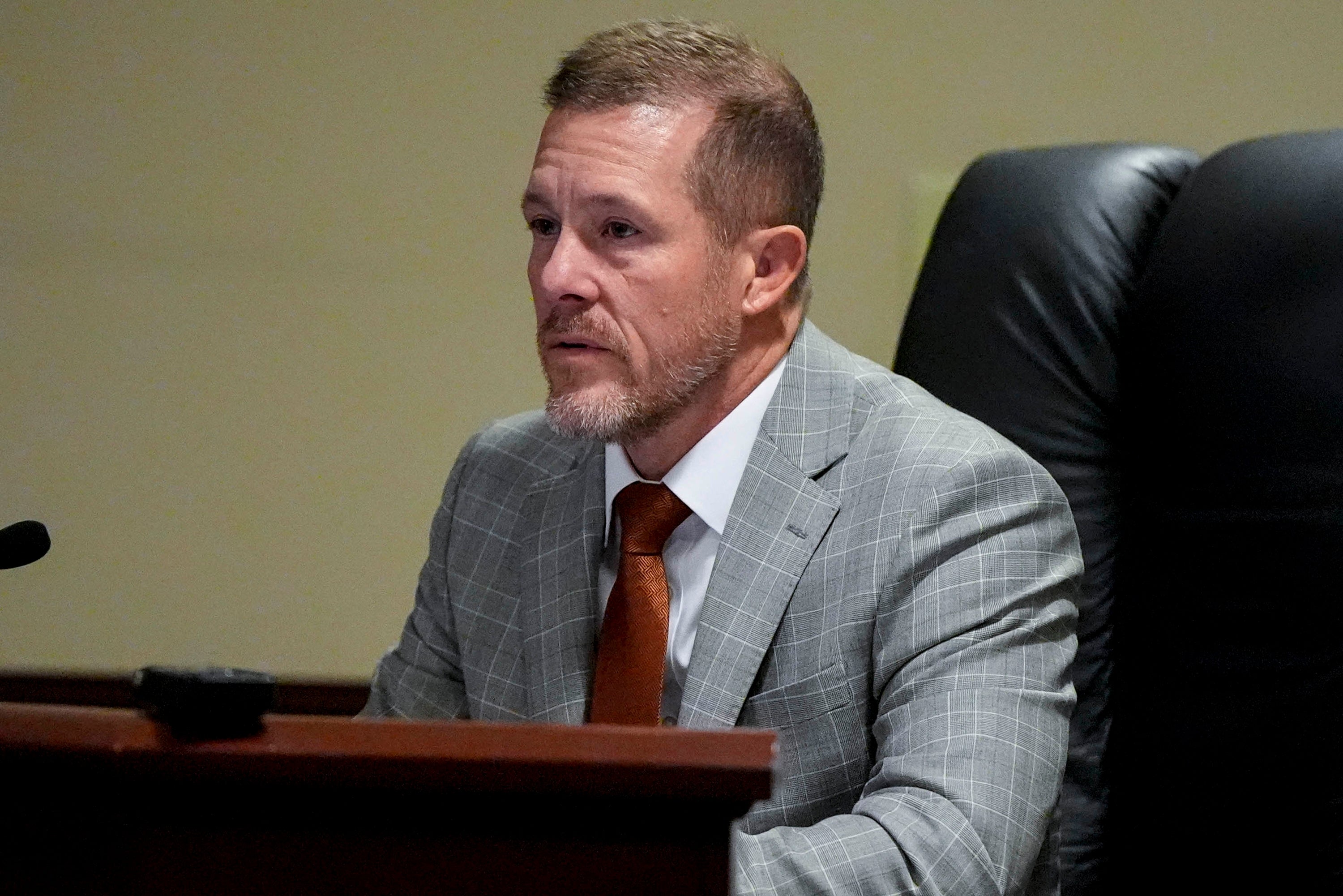 A man in a suit and tie speaks at a microphone, seated at a desk in a formal setting