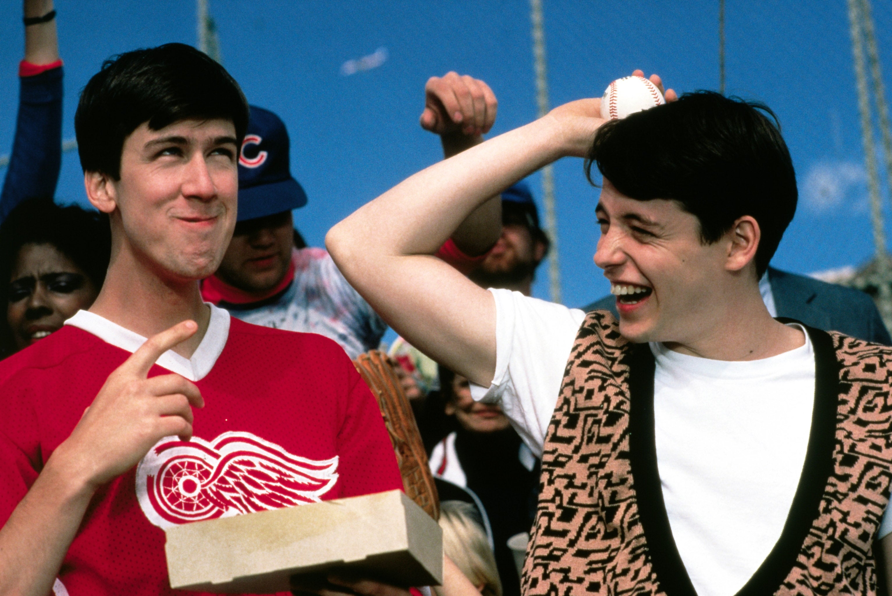 Two young men at a baseball game; one wears a sports jersey, the other a patterned sweater vest. They share a playful, cheerful moment