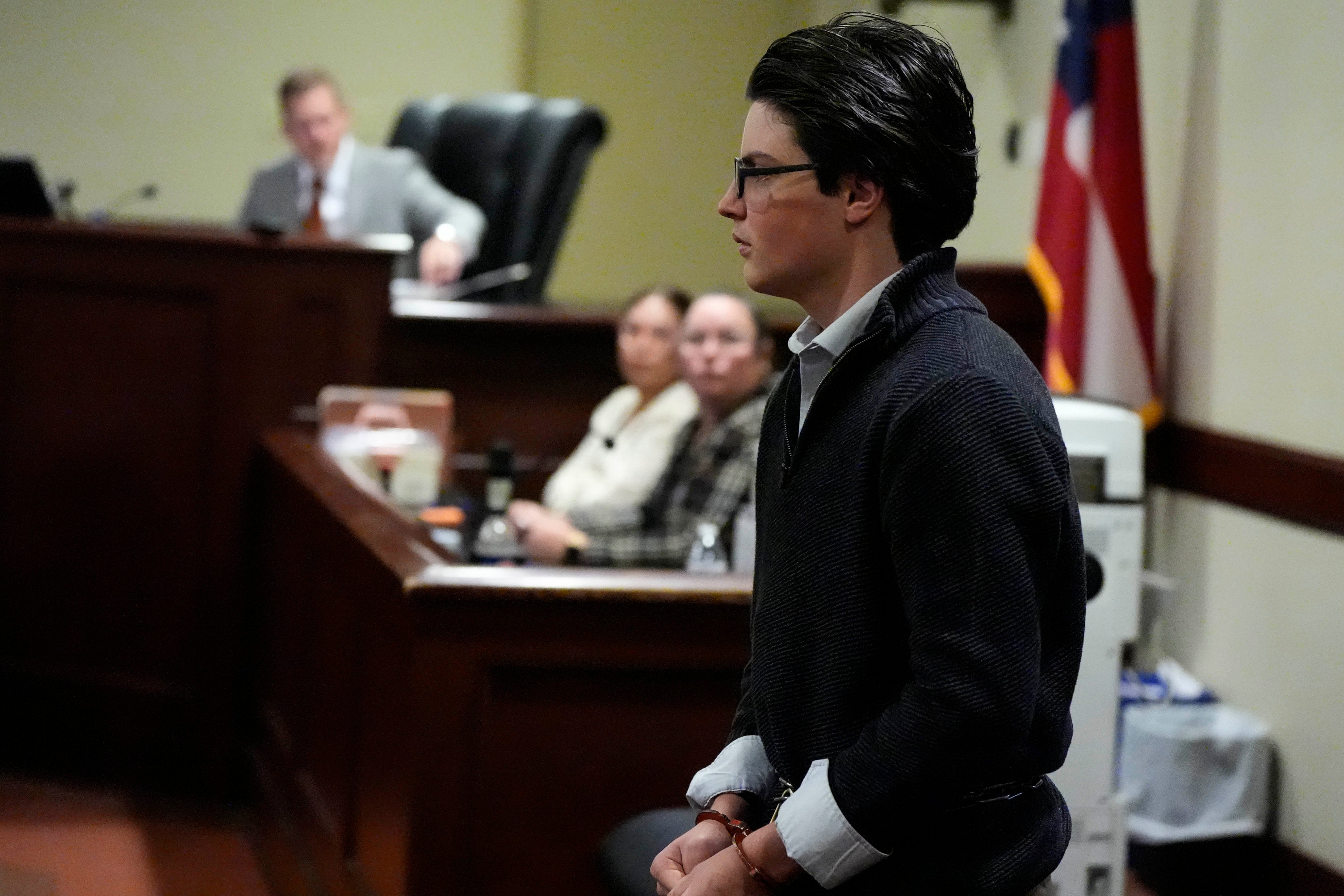 A person in a sweater and glasses stands in court, with people seated attentively in the background