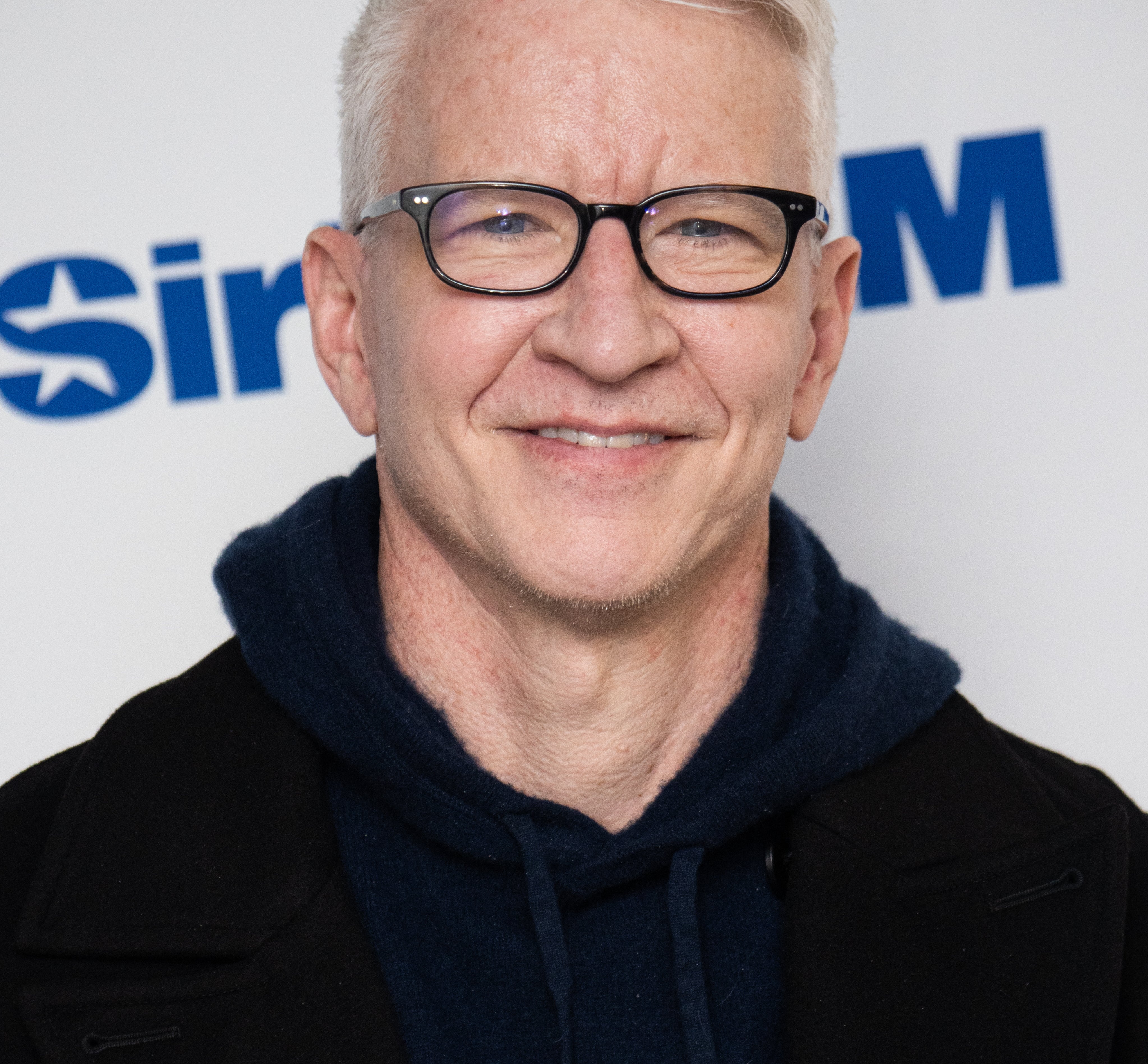 A person with short hair and glasses, wearing a dark hoodie and coat, smiling at a media event backdrop