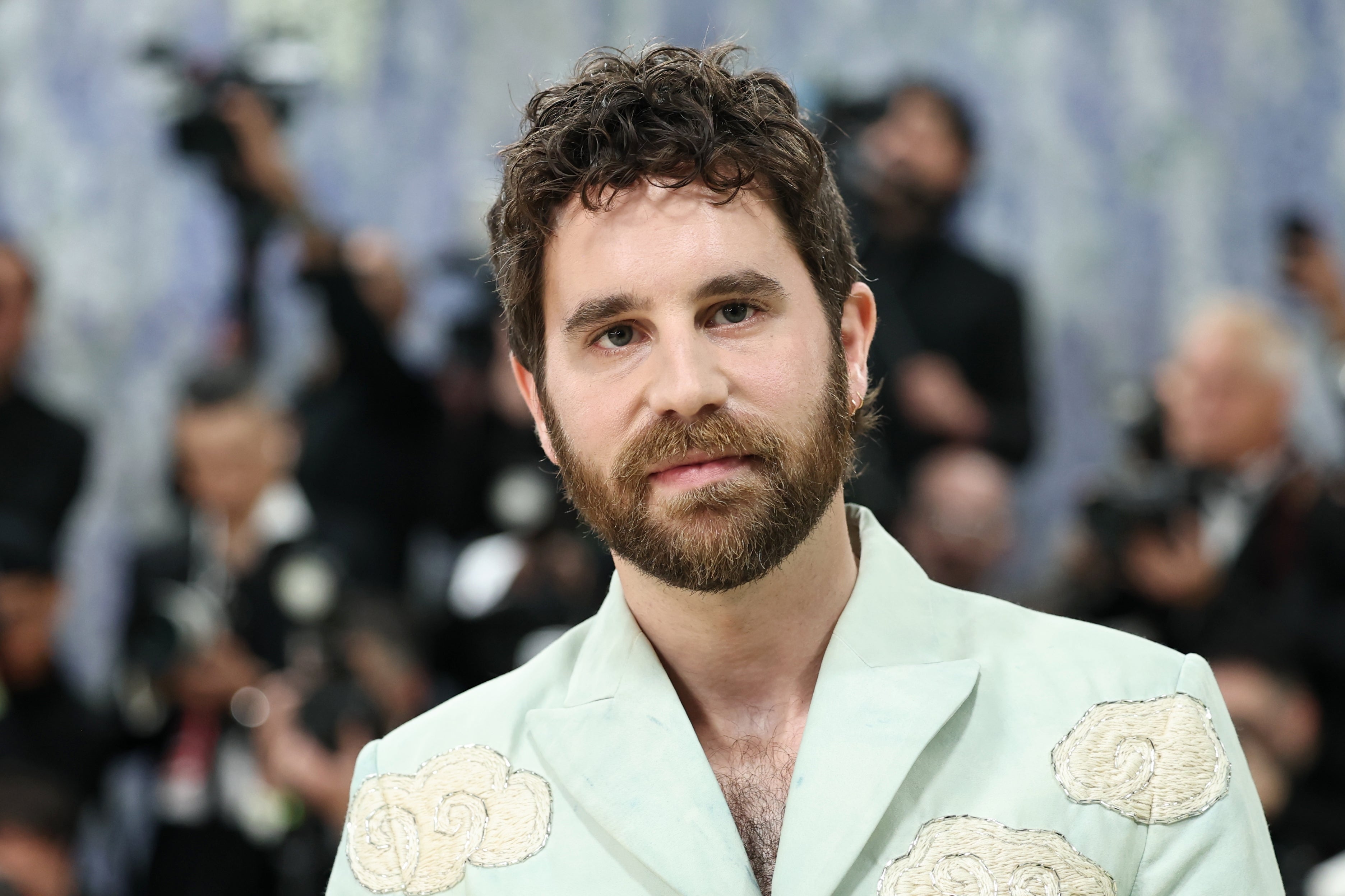 Individual on a red carpet wearing a tailored suit with decorative patterns, surrounded by photographers in the background