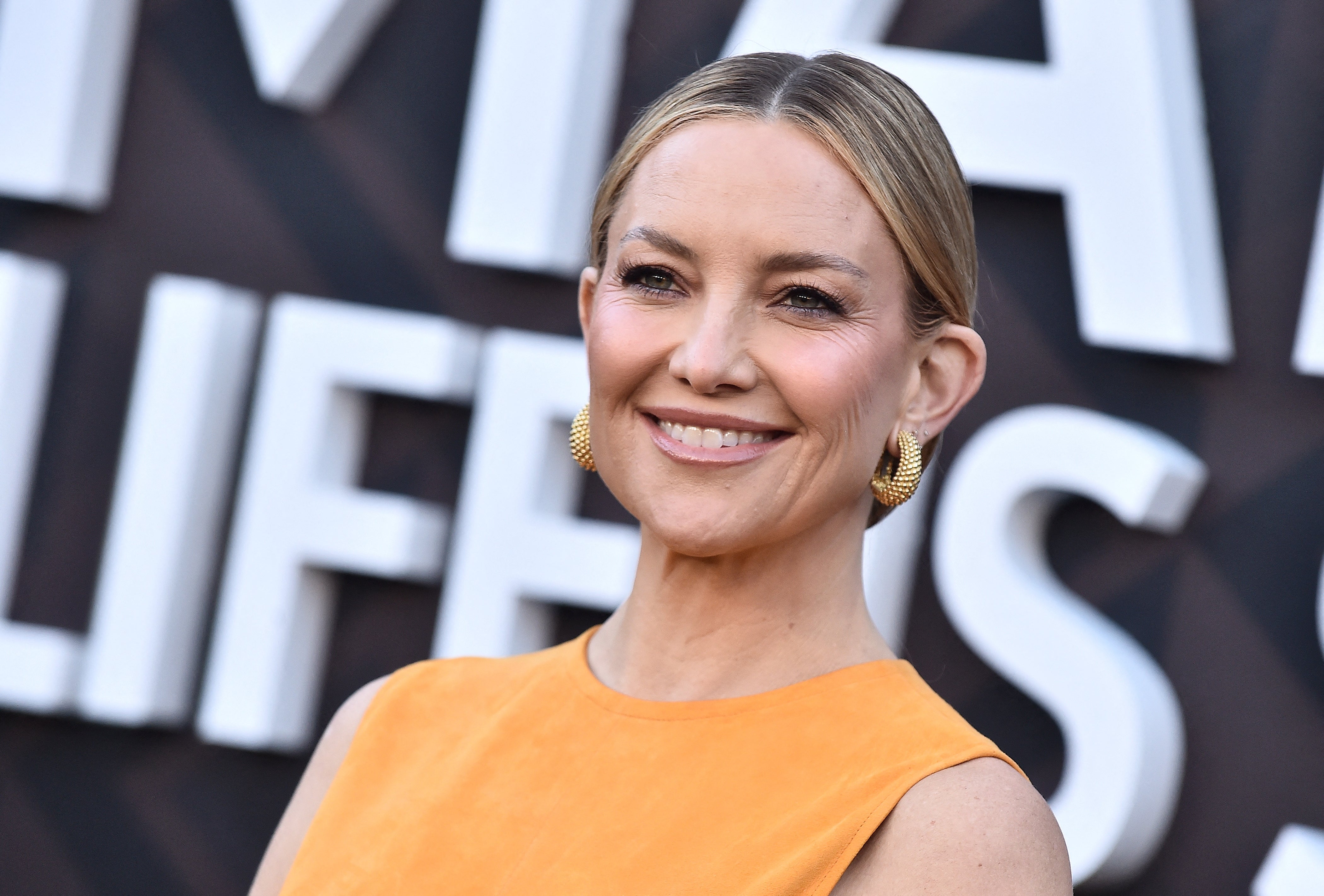 A person smiling on a red carpet, wearing a sleeveless outfit and gold hoop earrings, in front of a background with large text