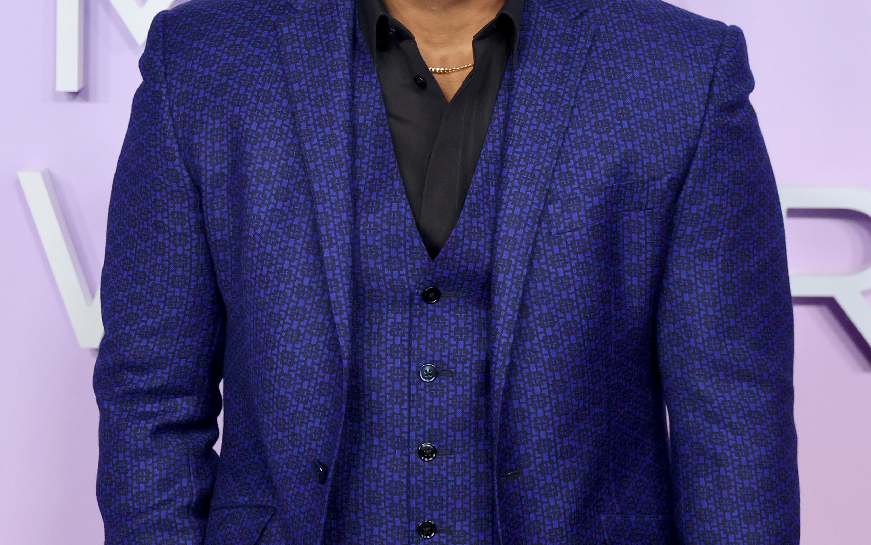 Man on red carpet in a tailored three-piece suit, wearing a black shirt