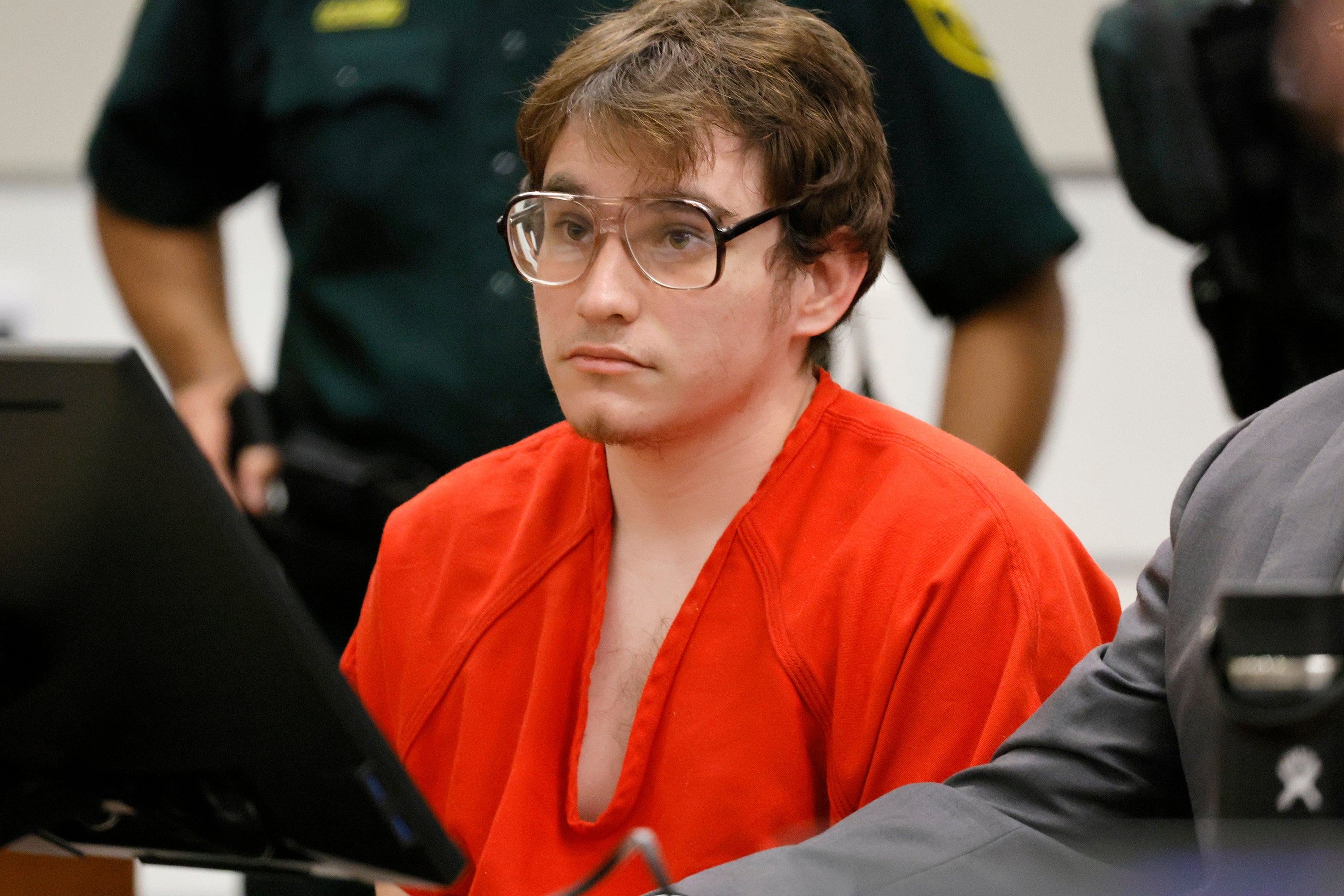 A person in a courtroom wearing glasses and a red jumpsuit sits at a table, appearing to listen intently
