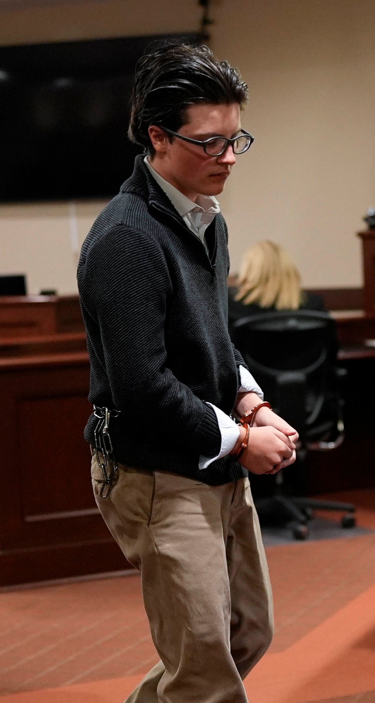 A person in handcuffs is escorted by two officers in a courtroom setting