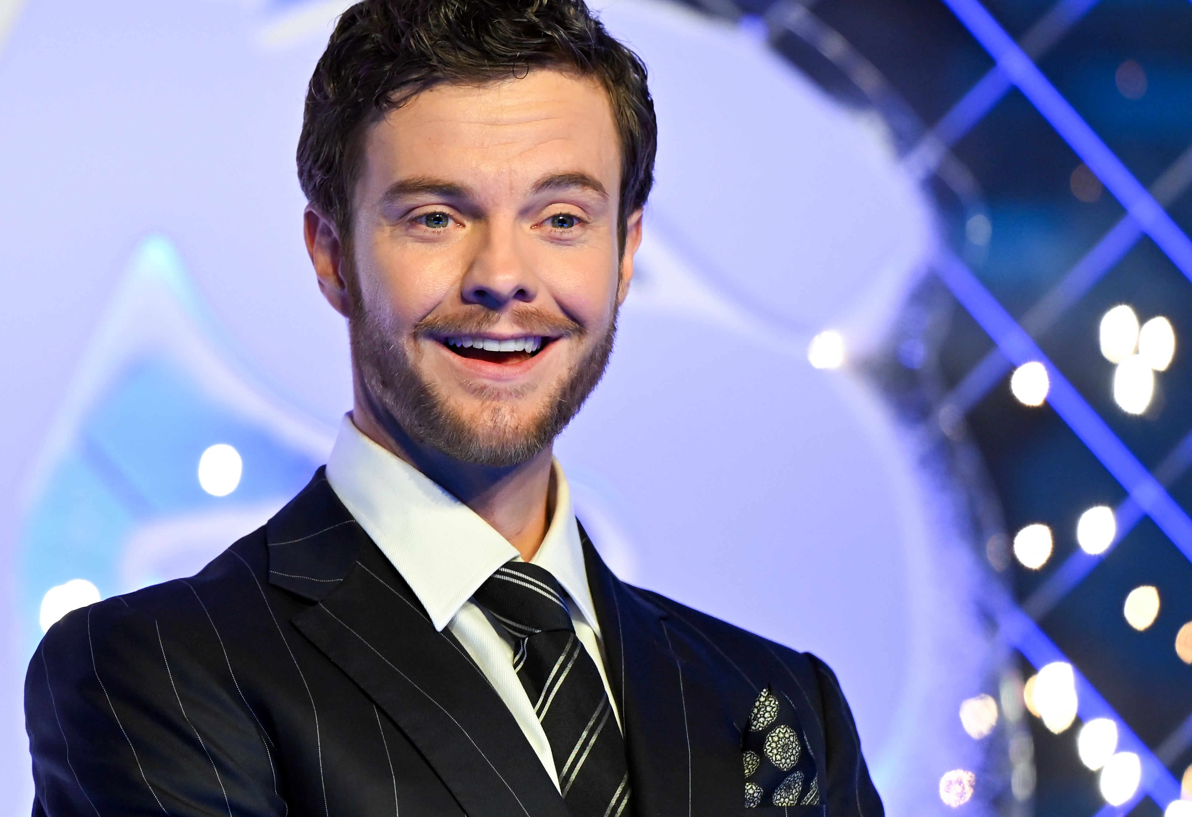 Person in pinstripe suit with tie smiles at an event, standing in front of a decorative backdrop with lights