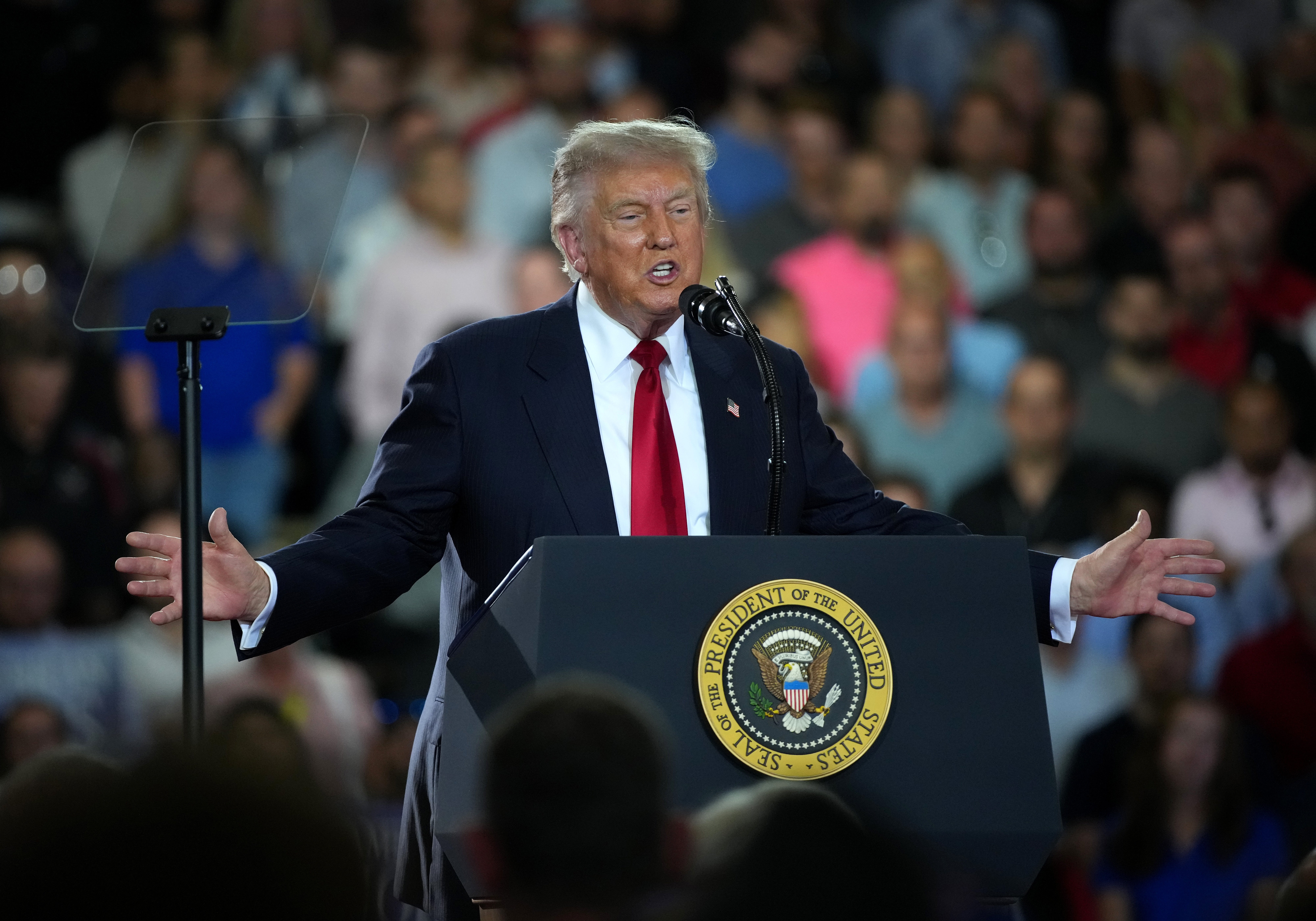 En mann i dress taler ved et podium med den amerikanske presidentens segl og holder armene vidt åpne for et publikum