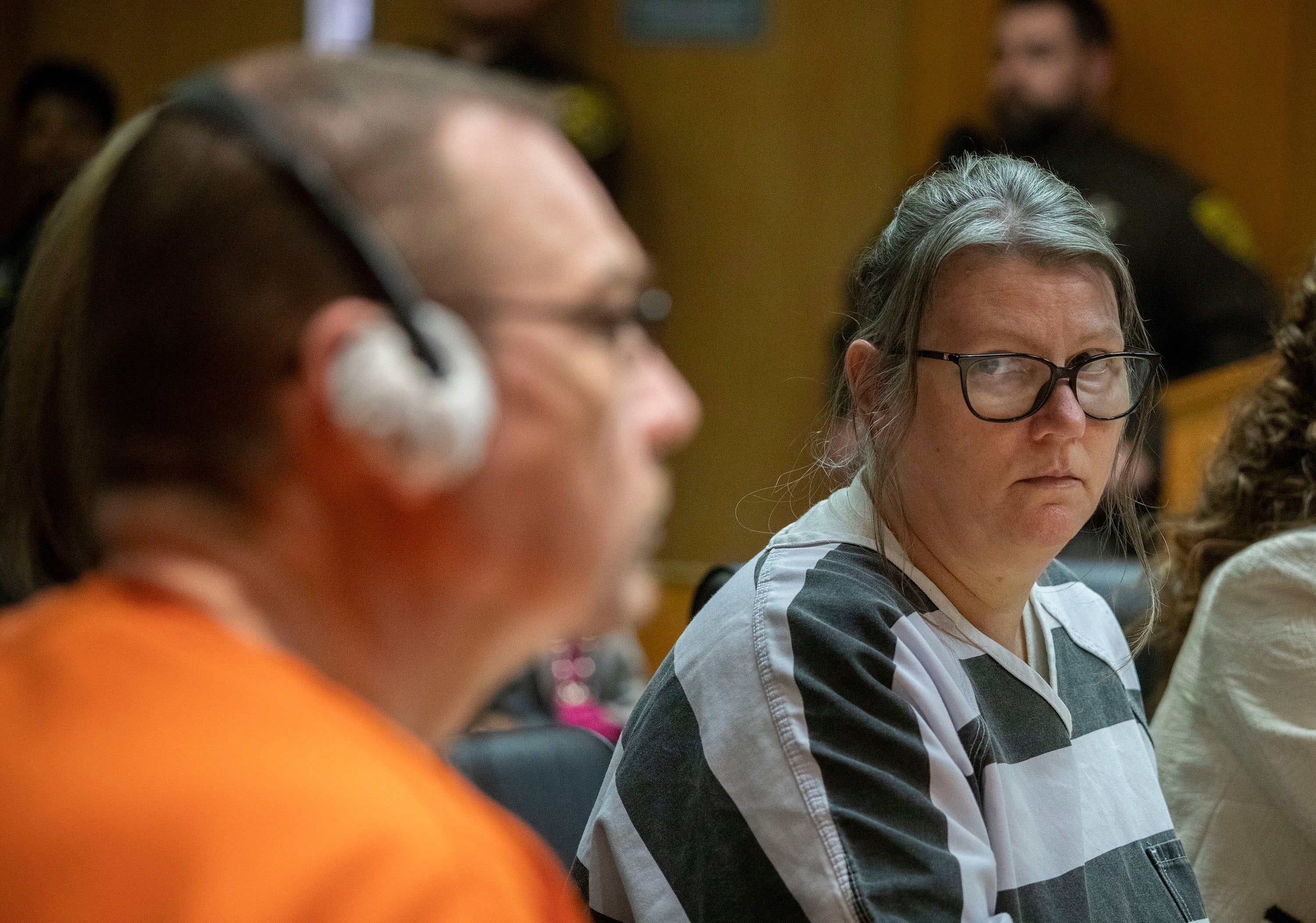 A woman in a striped prison jumpsuit sits in a courtroom, looking towards a man in an orange jumpsuit