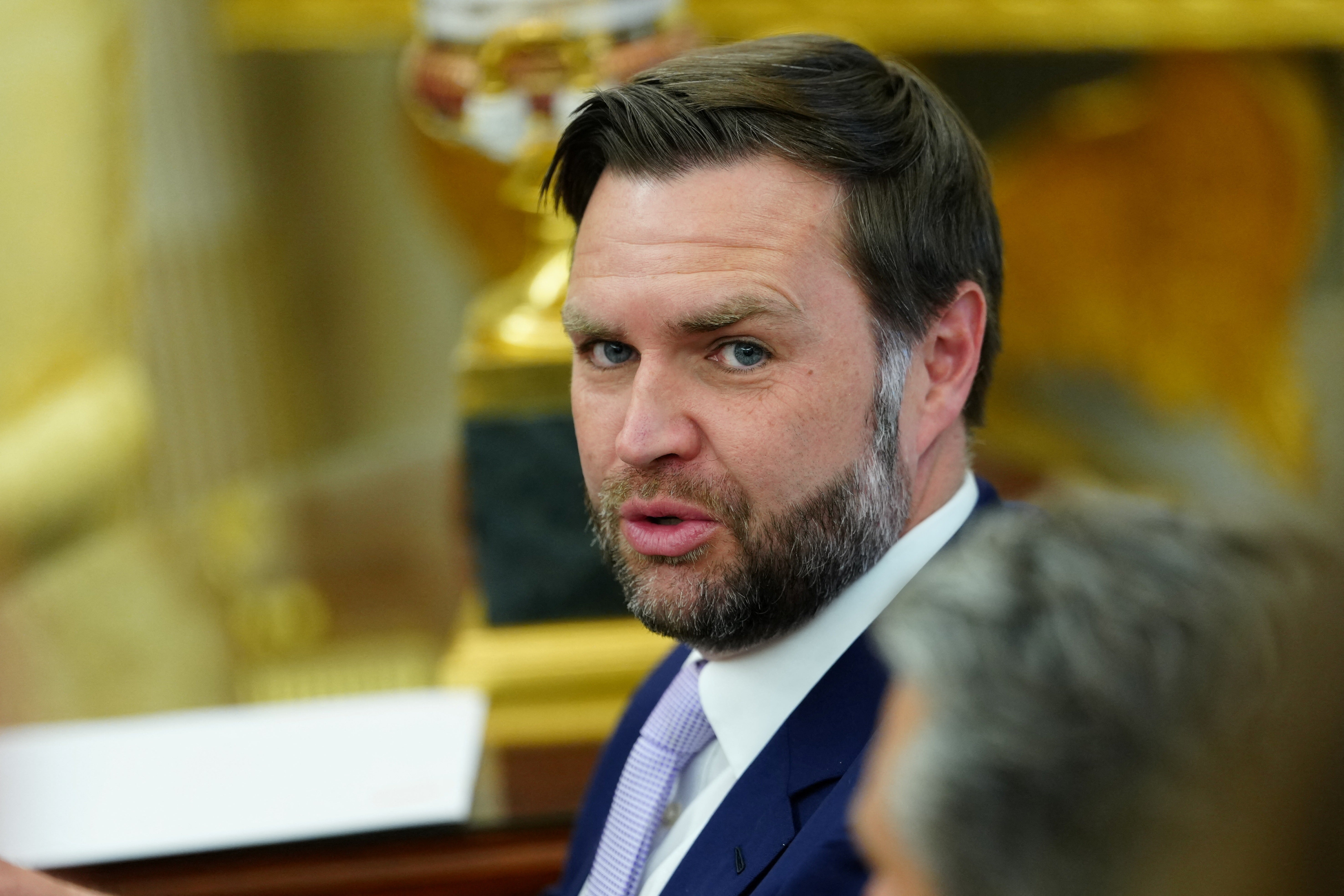 Mann i dress og slips snakker, sittende innendørs med utsmykket dekor i bakgrunnen