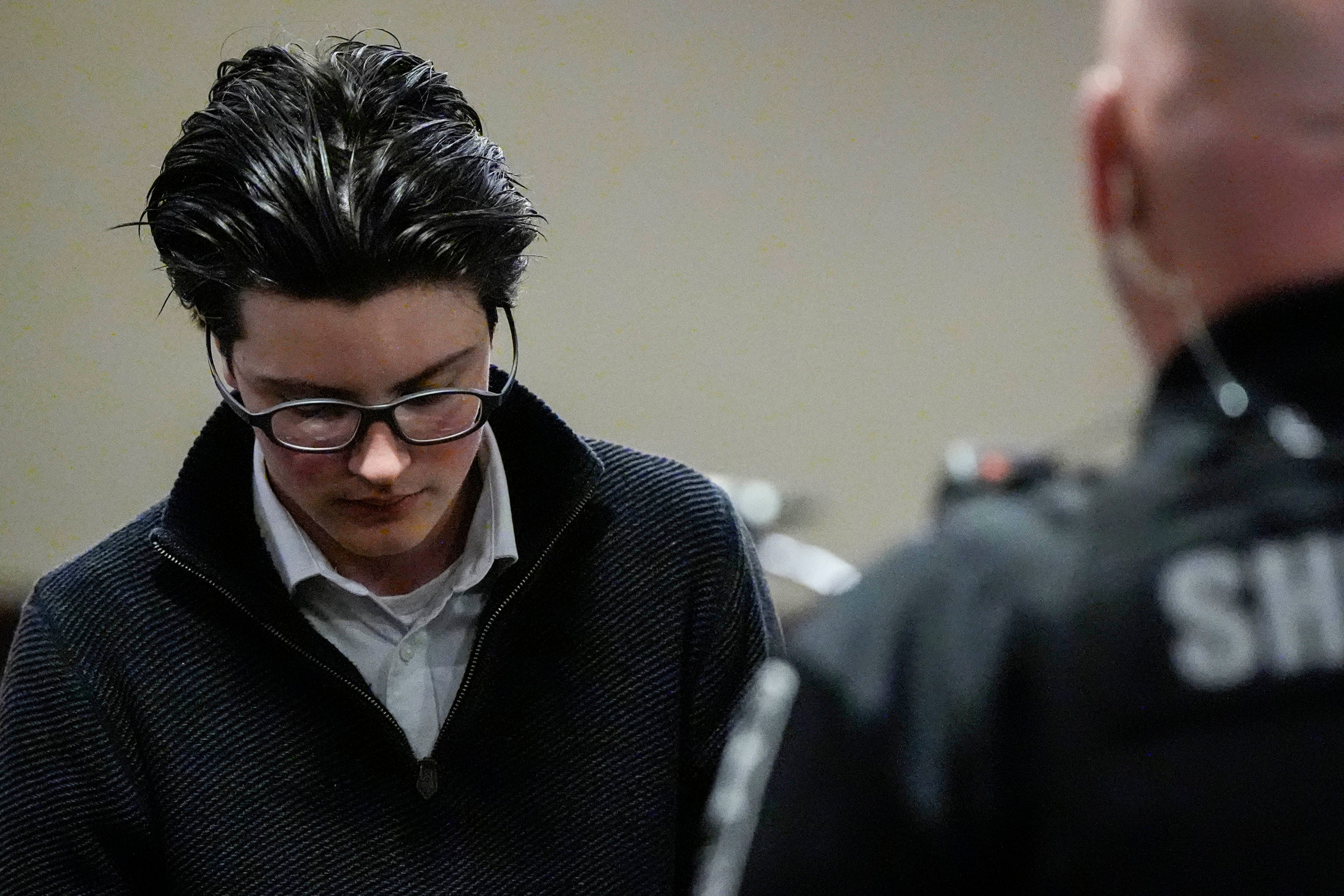 Person with glasses and dark hair, wearing a sweater over a collared shirt, stands in a room with a blurred uniformed officer in the foreground