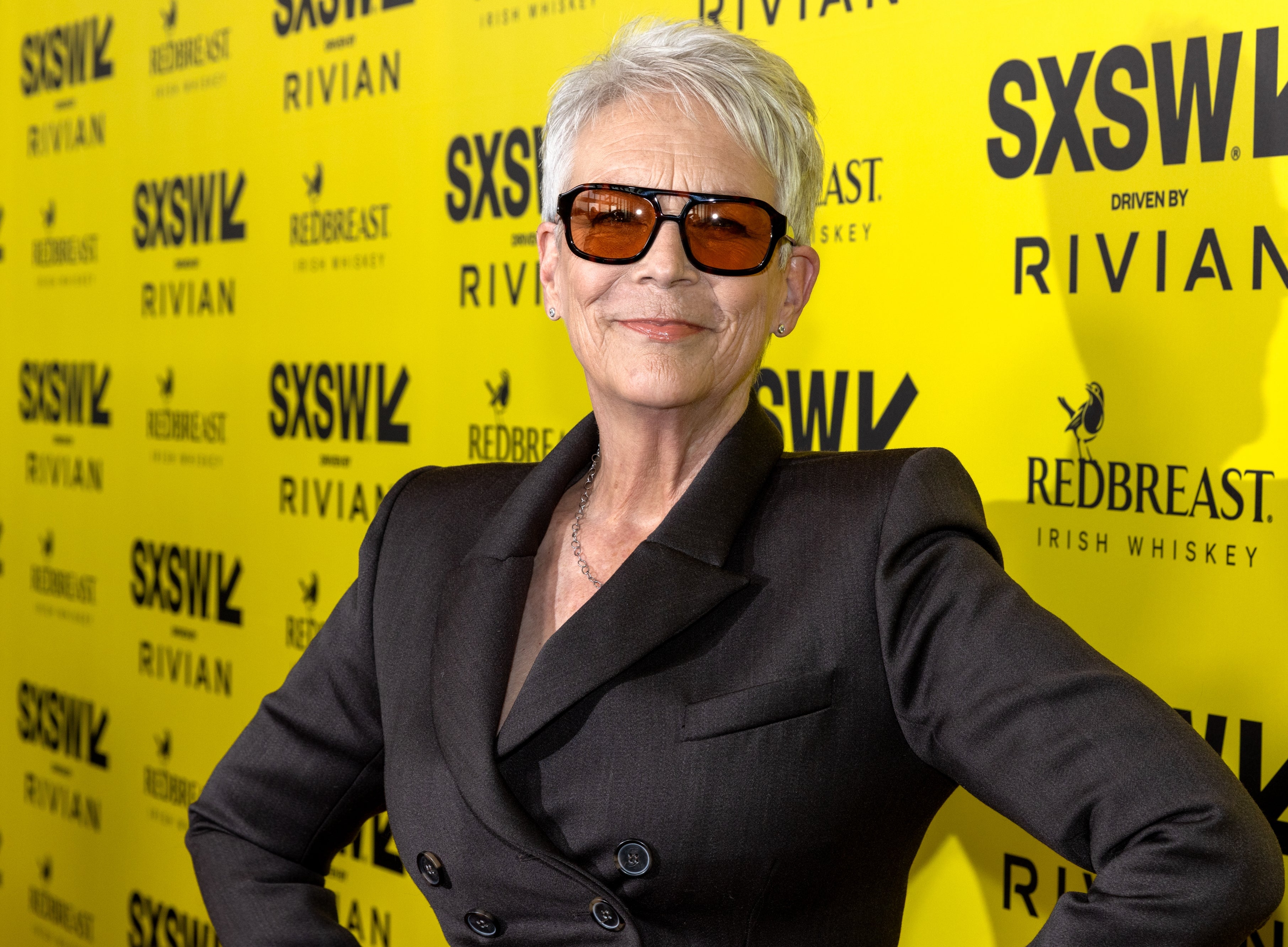 Person in sleek suit with sunglasses poses confidently at SXSW red carpet event with branded background