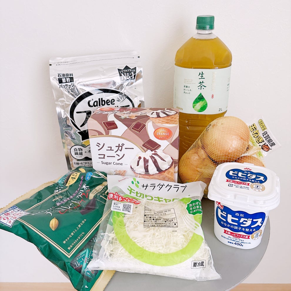 KALDI（カルディ）の「コンパクト保冷バッグ ブラック 1枚」テーブルの上に千切りキャベツ、玉ねぎ、お茶、ヨーグルト、グラノーラ、アイス、チョコレートが並んでいる。