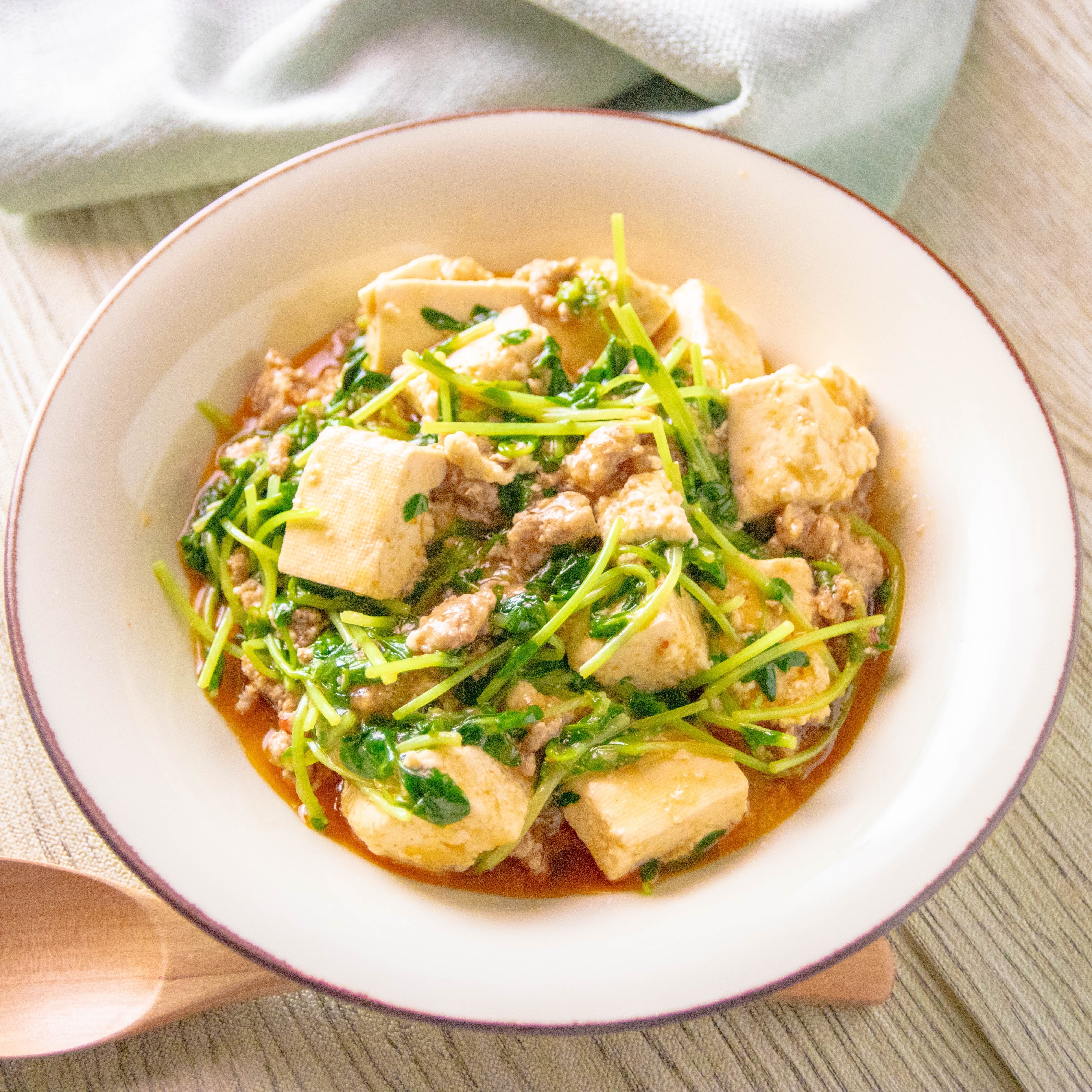 深皿に盛られた豆苗と豆腐のピリ辛あん。豚ひき肉が入り、木製スプーンが添えられています。