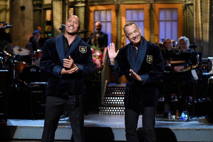 Two unidentified men in matching dark robes with number 5 emblems, smiling on stage during a live performance