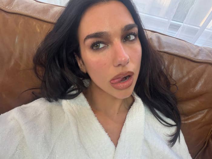 A person with long dark hair, wearing a white robe, poses on a leather couch with a relaxed expression and slightly open mouth
