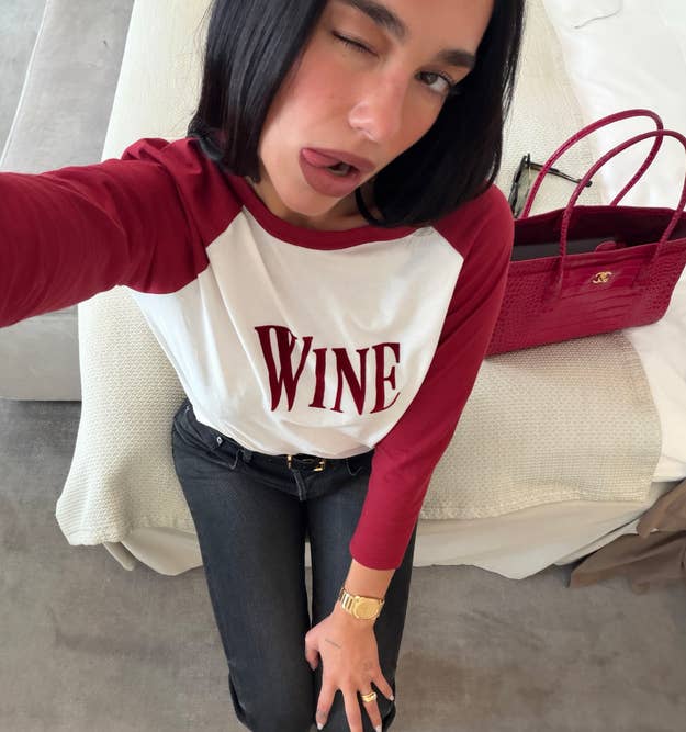 Person winking, wearing a shirt with "WINE" on it, casual style, posing near a red handbag on a bed