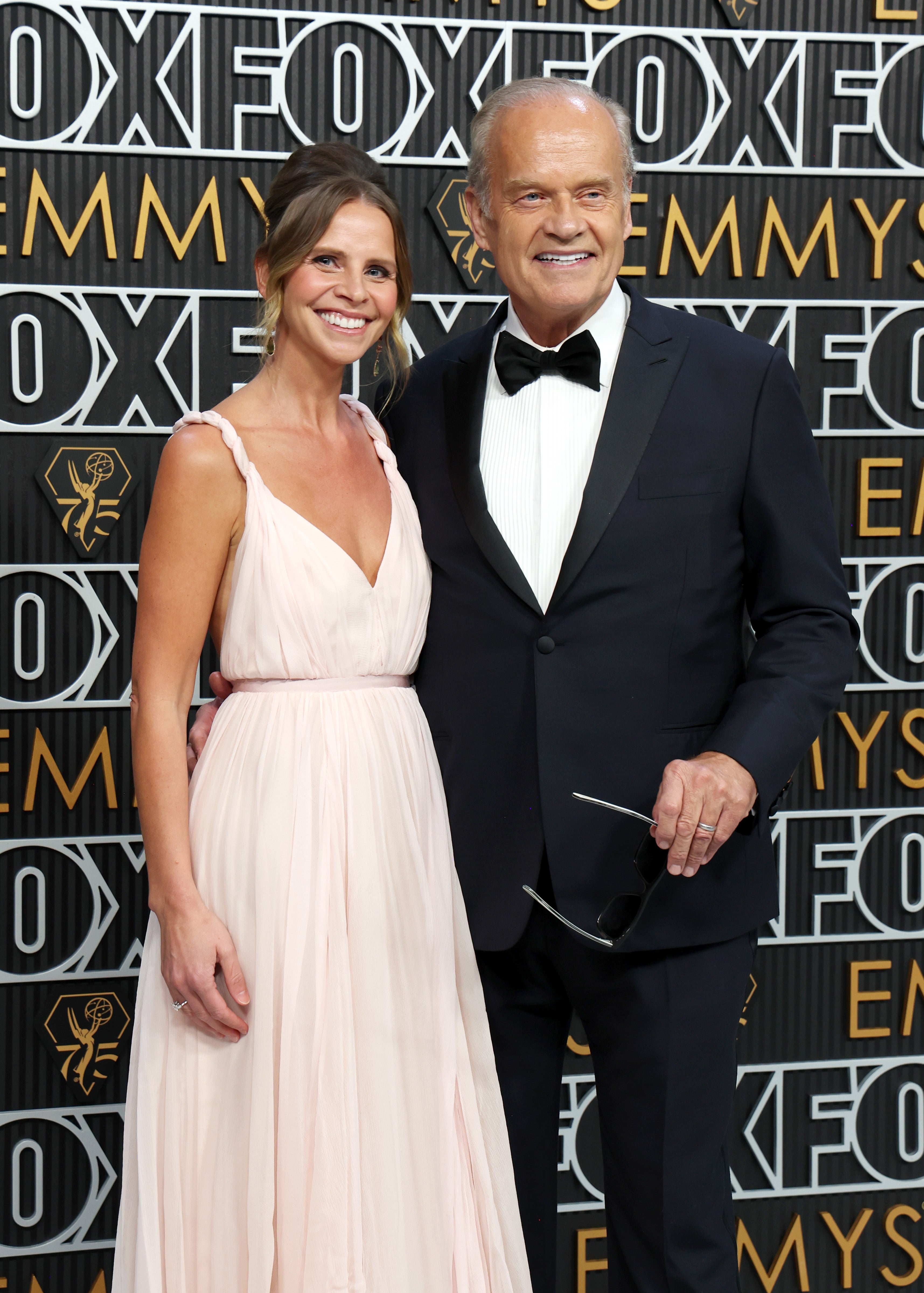 Two people posing on a red carpet. The man wears a tuxedo, and the woman wears an elegant, flowing dress