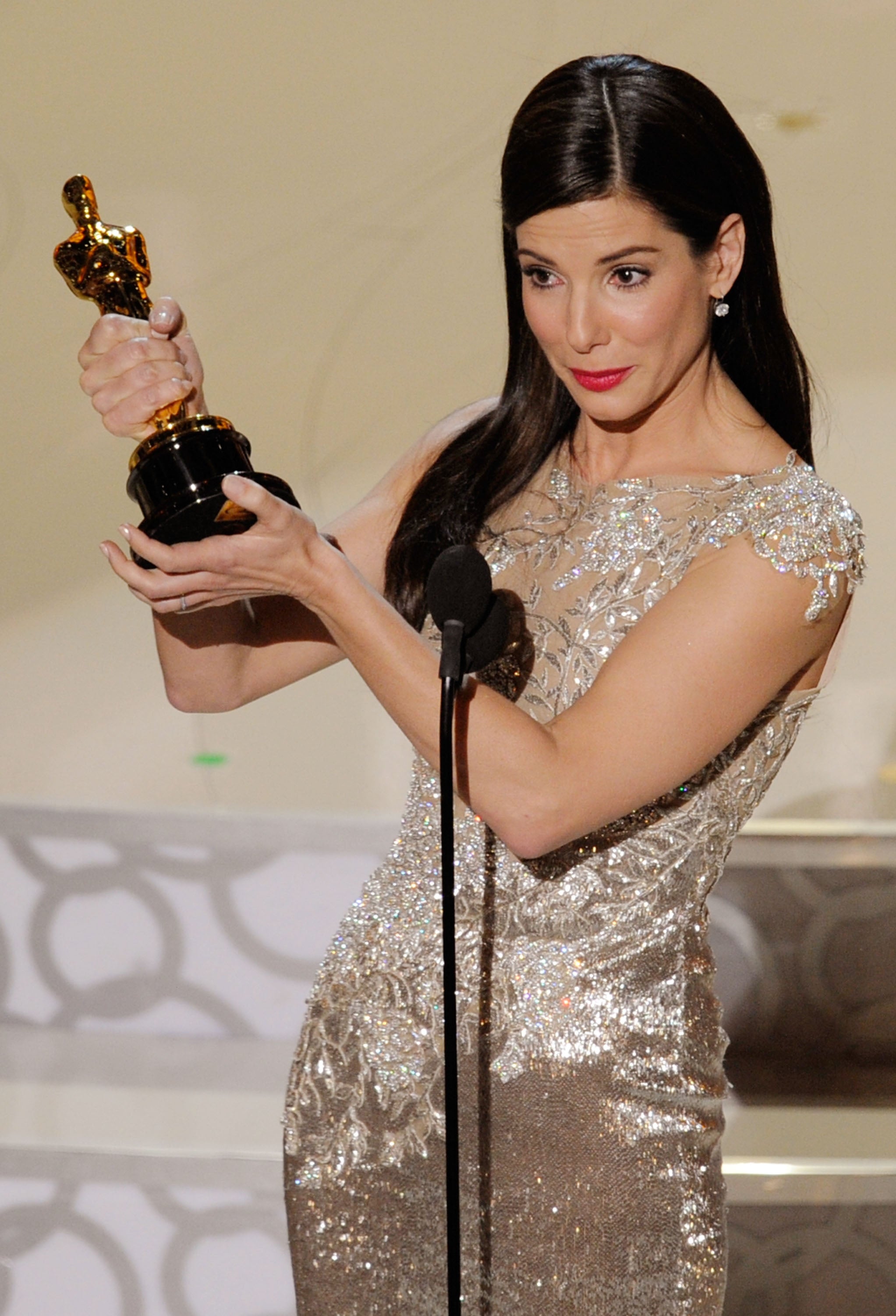 Celebrity in an elegant gown holds an award while speaking at a podium