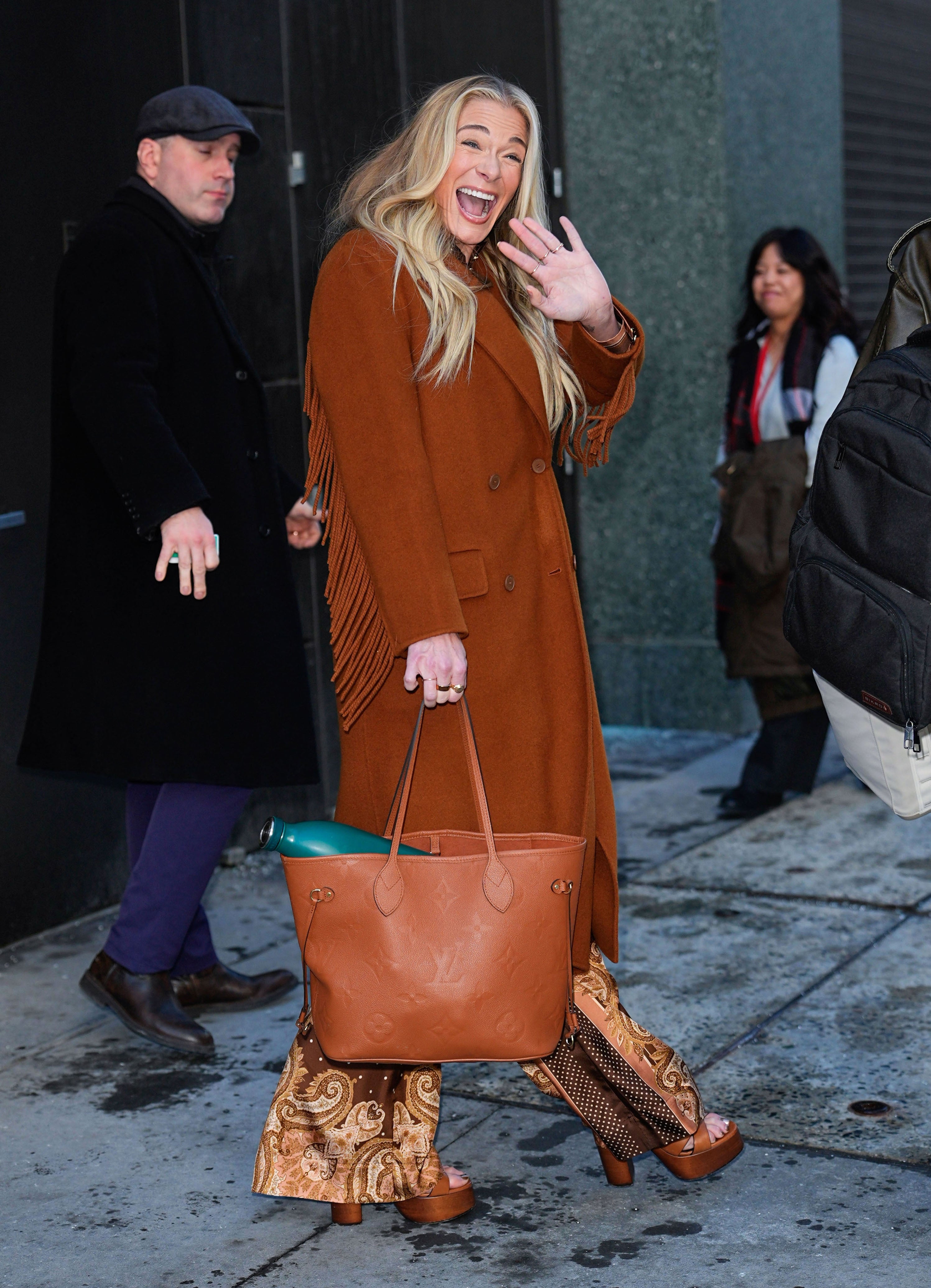 Celebrity waving and smiling, wearing a long brown coat with fringe details and patterned flared pants, holding a large tote bag