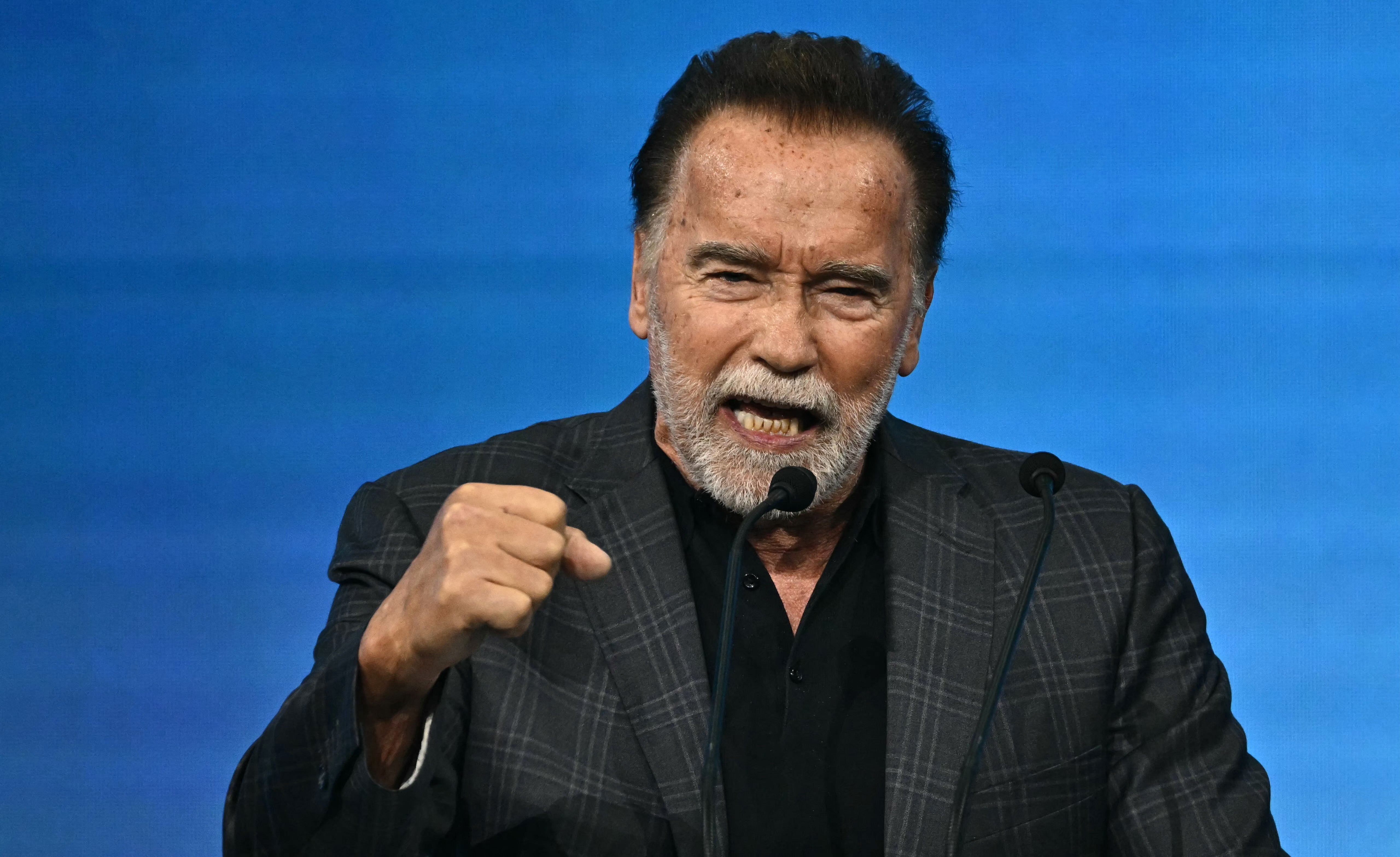 A public figure passionately speaking at an event, wearing a checkered suit jacket and solid shirt, with a raised fist gesture