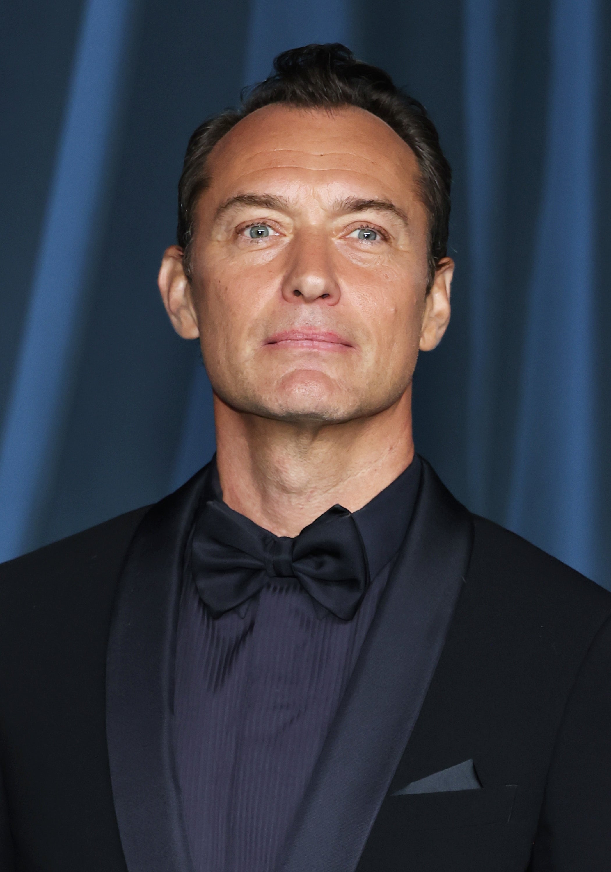 Person in formal wear with a dark tuxedo and bow tie, posing against a draped background