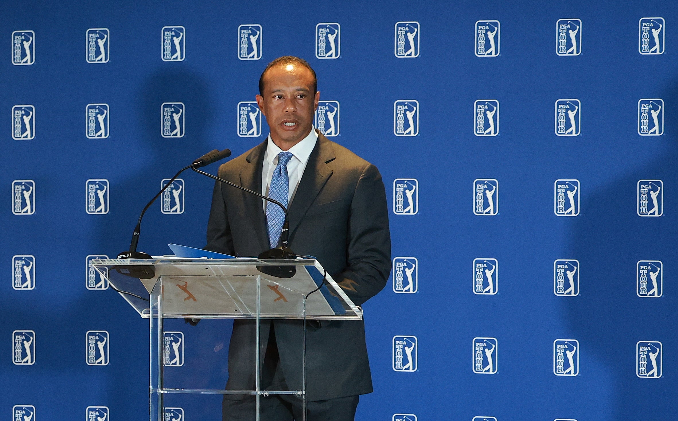 A person in a suit speaks at a podium during a PGA event, with logo backdrop