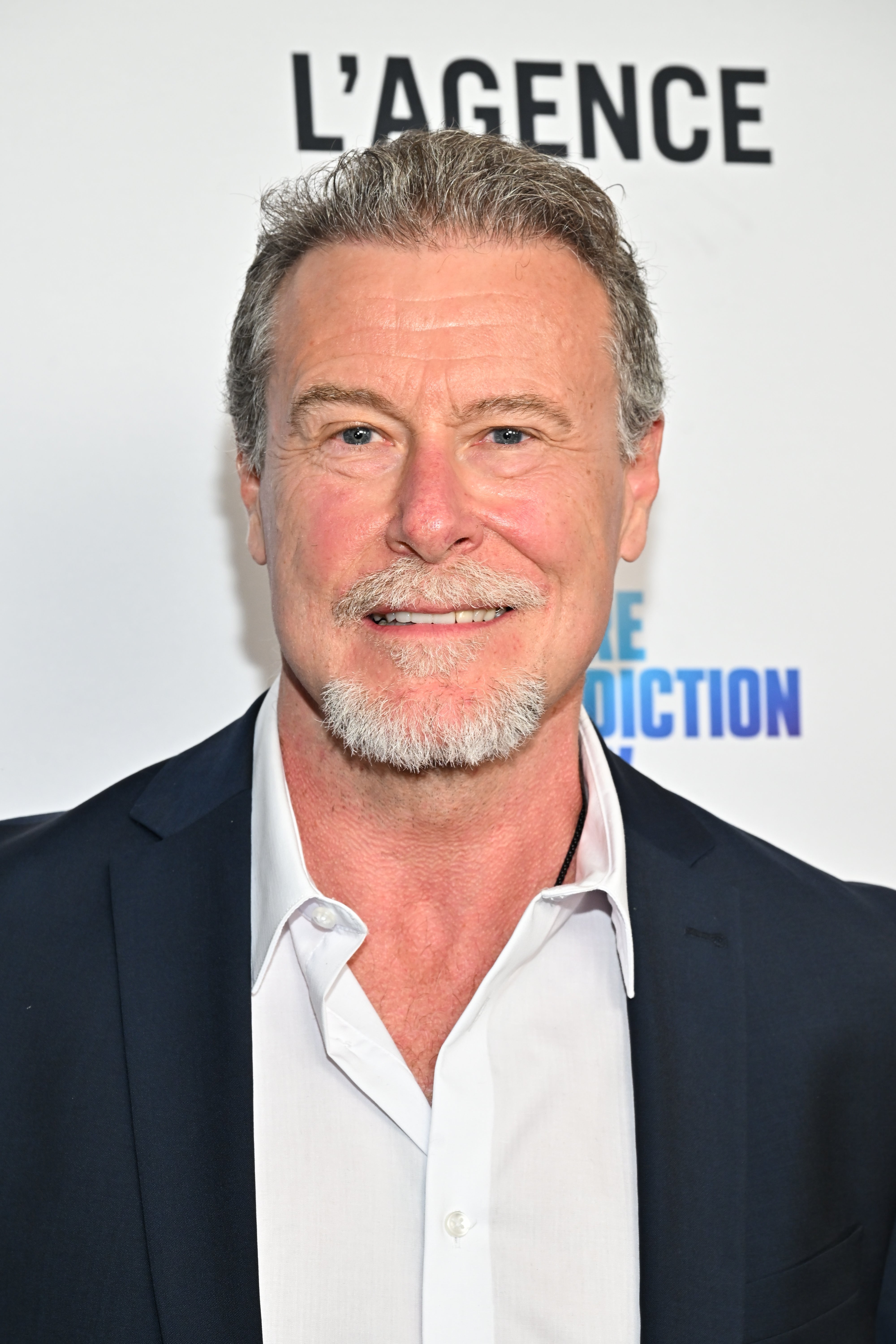 Smiling man with short hair and a goatee in a white shirt and dark blazer at a celebrity event backdrop