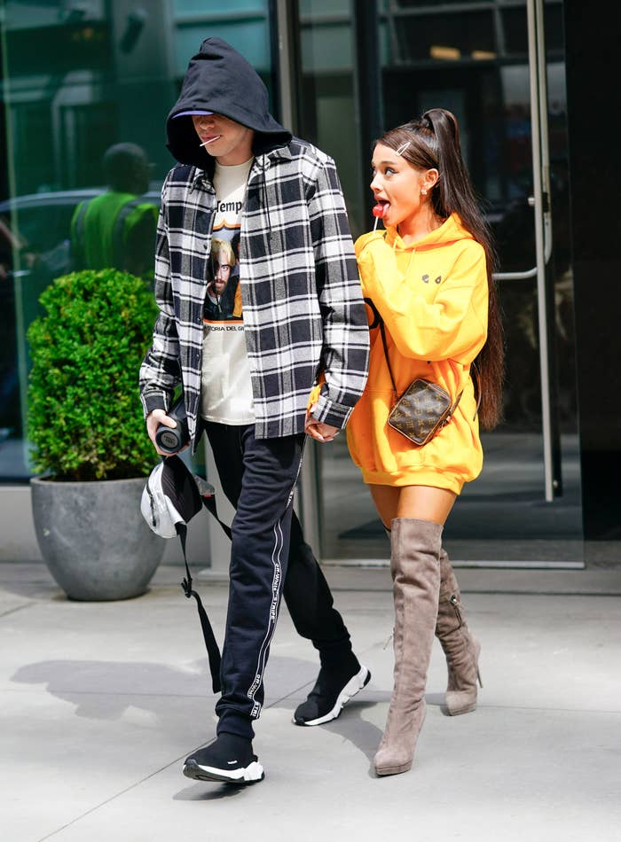 Two people walk hand-in-hand. One wears a plaid shirt with a hood and sneakers. The other sports an oversized hoodie, long boots, and a small crossbody bag