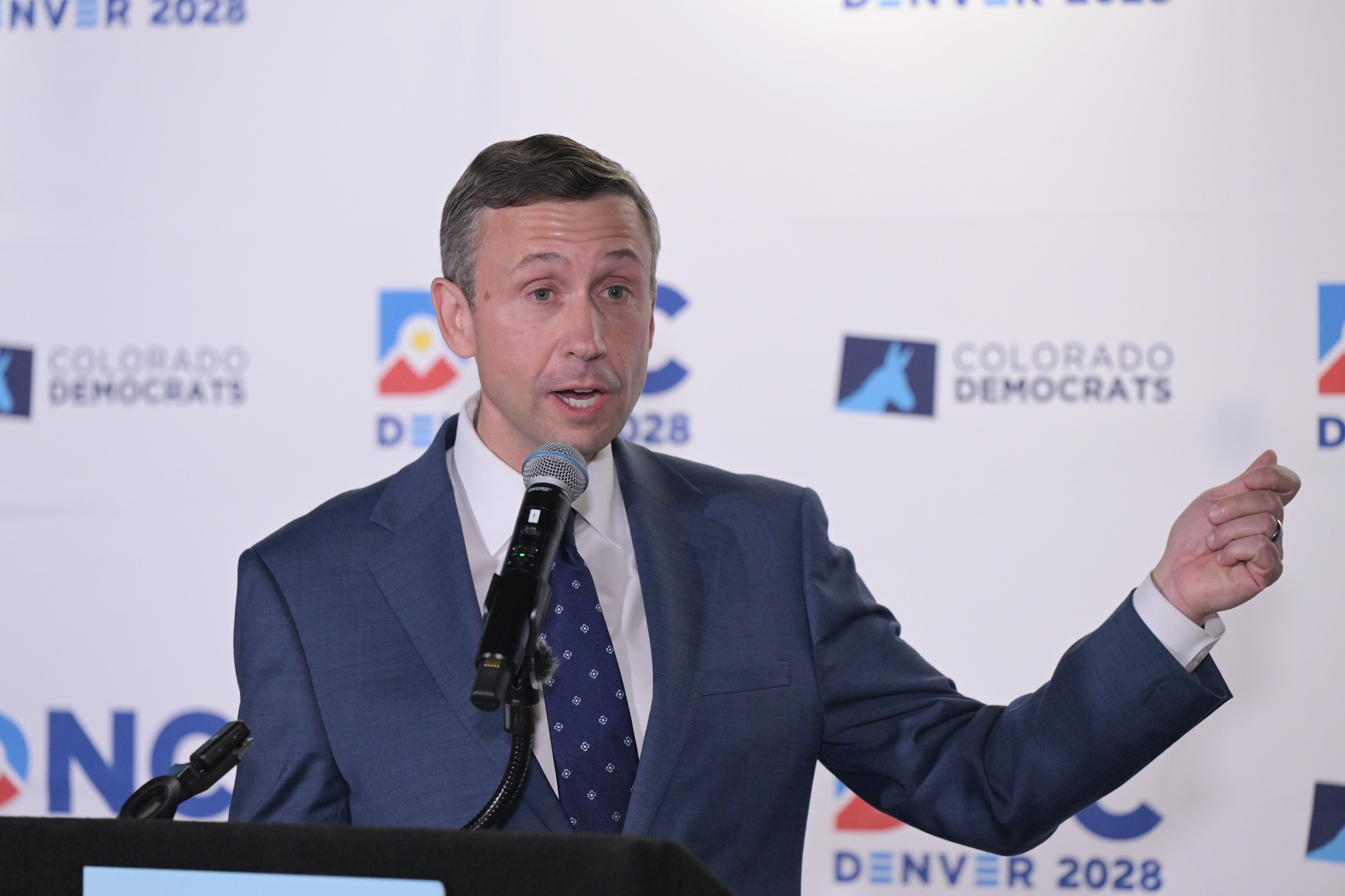 En mann i dress snakker ved et podium under en pressebegivenhet for DNC Denver 2028, med logoer i bakgrunnen
