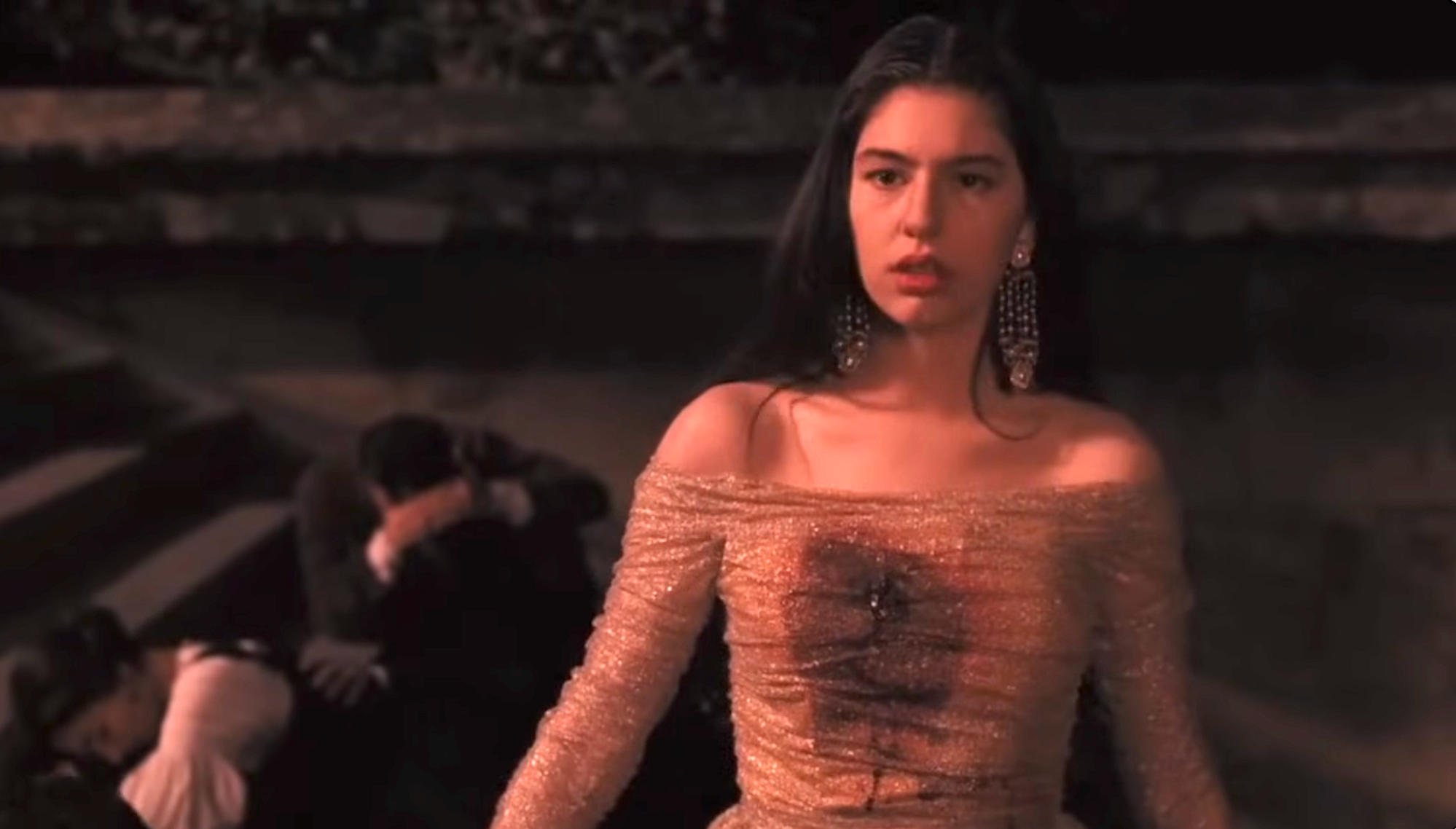 Sofia Coppola, in an elegant, off-shoulder gown, appears serious as she has just been shot in the chest. People in the background seem distressed and duck on the ground