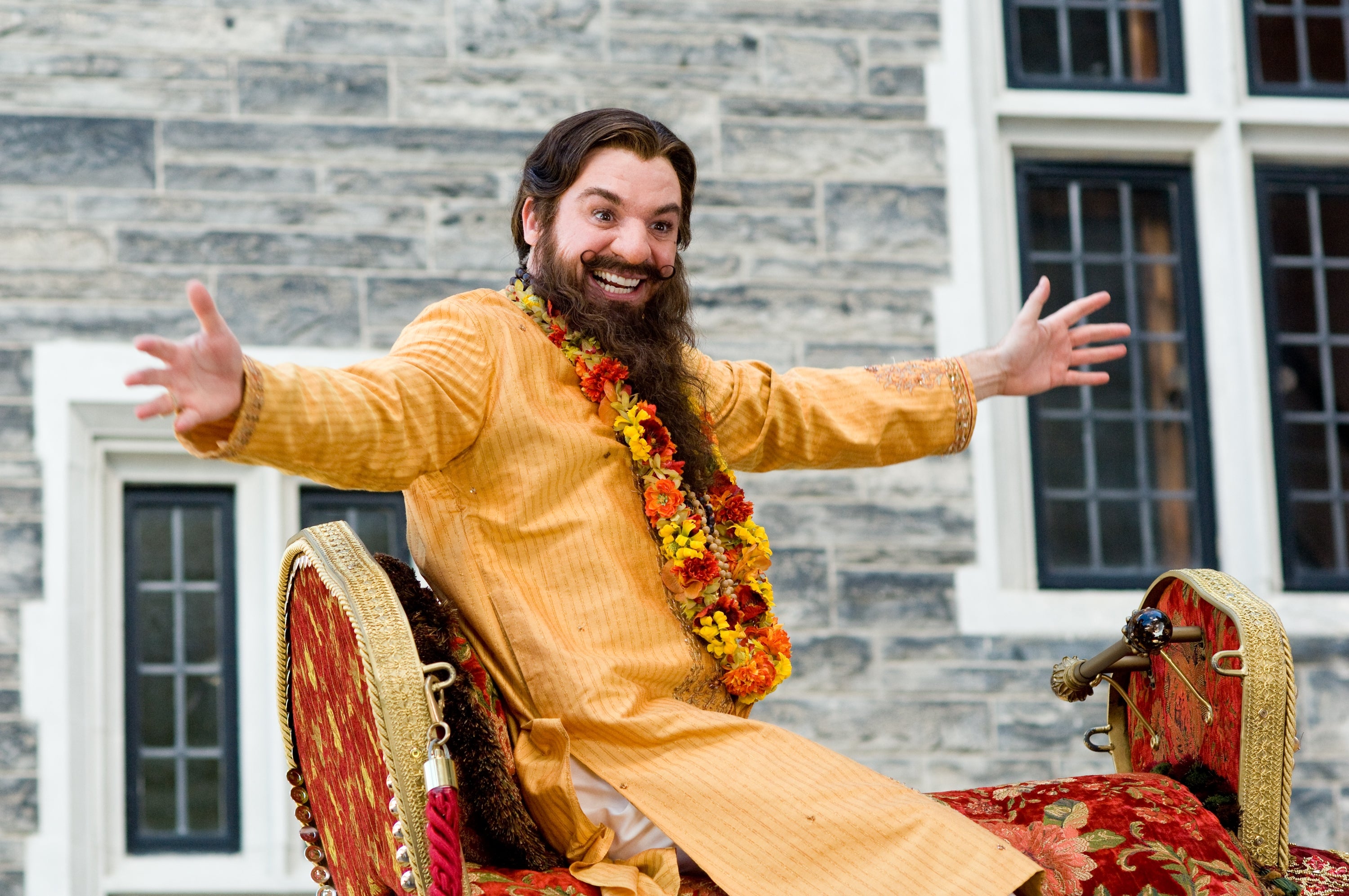Mike Myers, wearing a traditional outfit and a floral garland, smiling and posing seated on an ornate chair