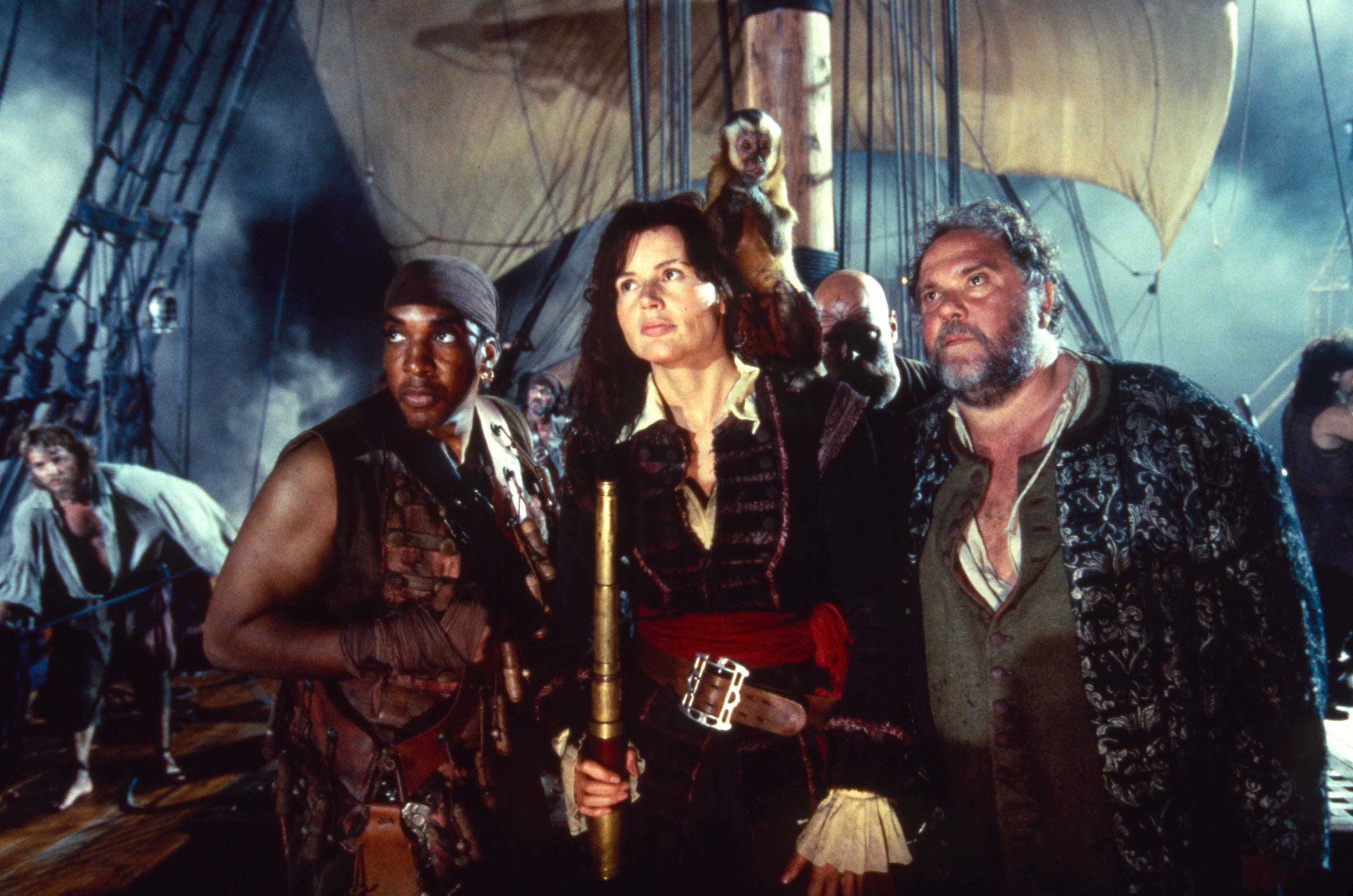 Geena Davis and others in pirate costumes stand dramatically on a ship's deck, holding a telescope