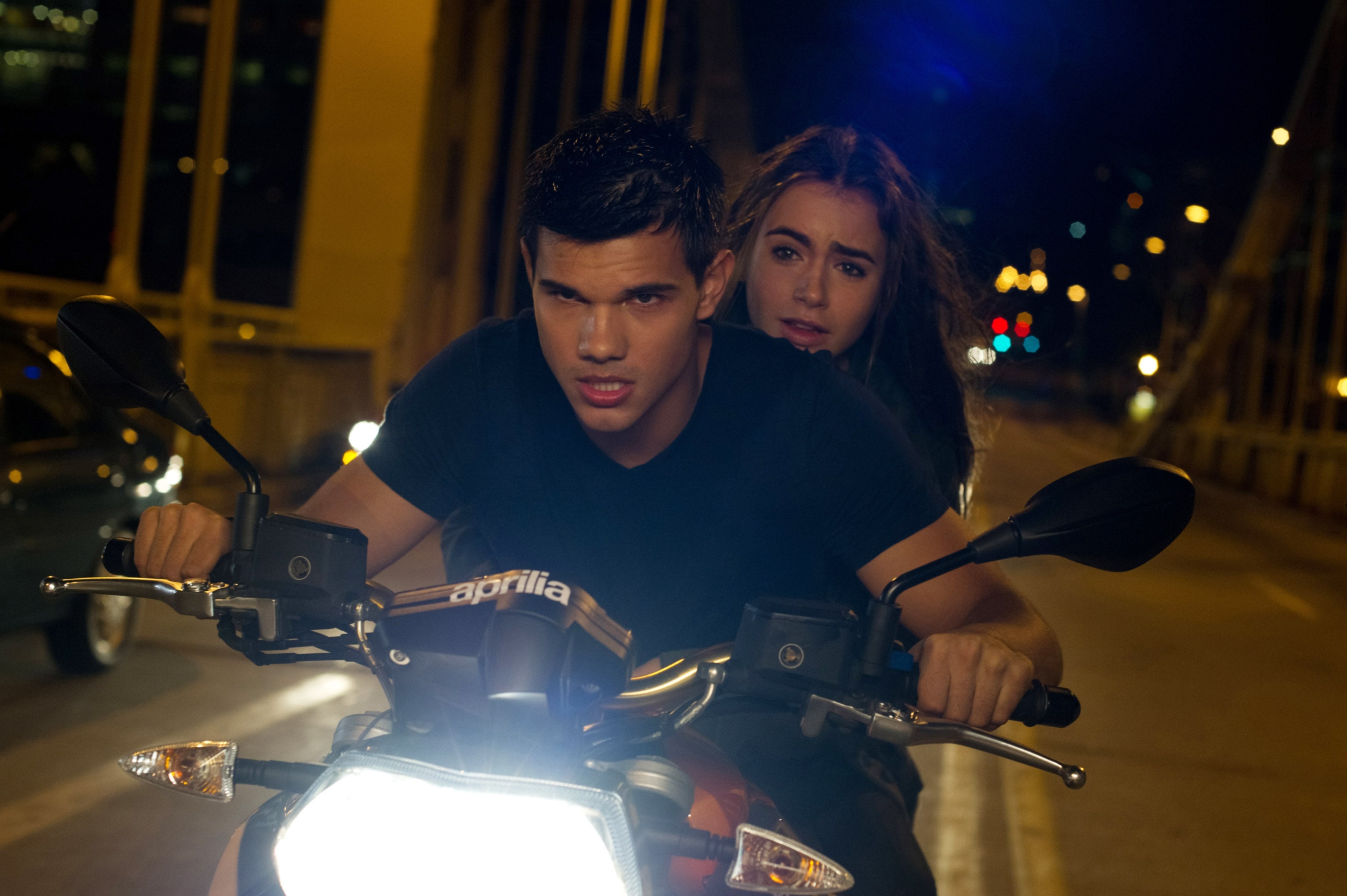 Taylor Lautner and Lily Collins ride a motorcycle on a bridge at night. The man in front is focused, and the woman behind him looks concerned