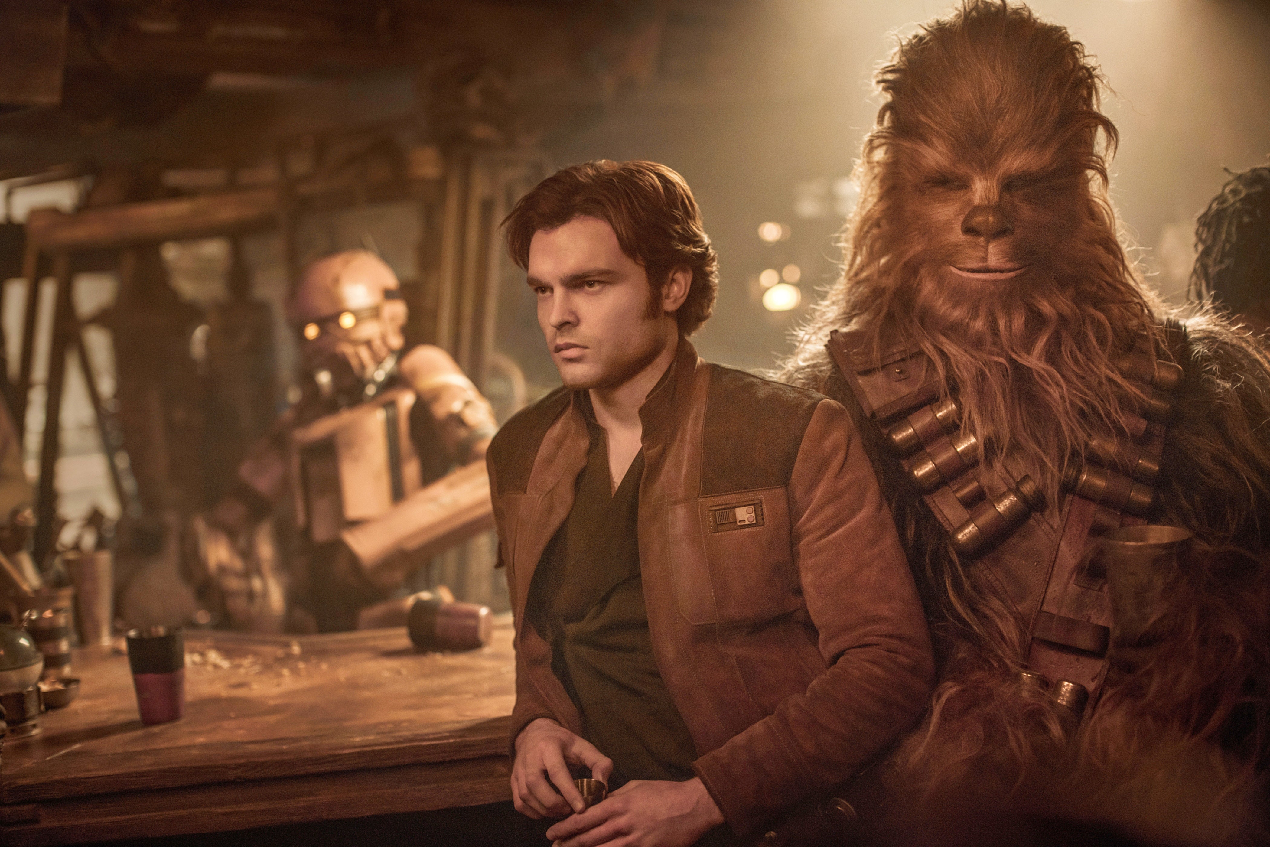 Alden Ehrenreich and Chewbacca stand in a dimly lit interior against a table