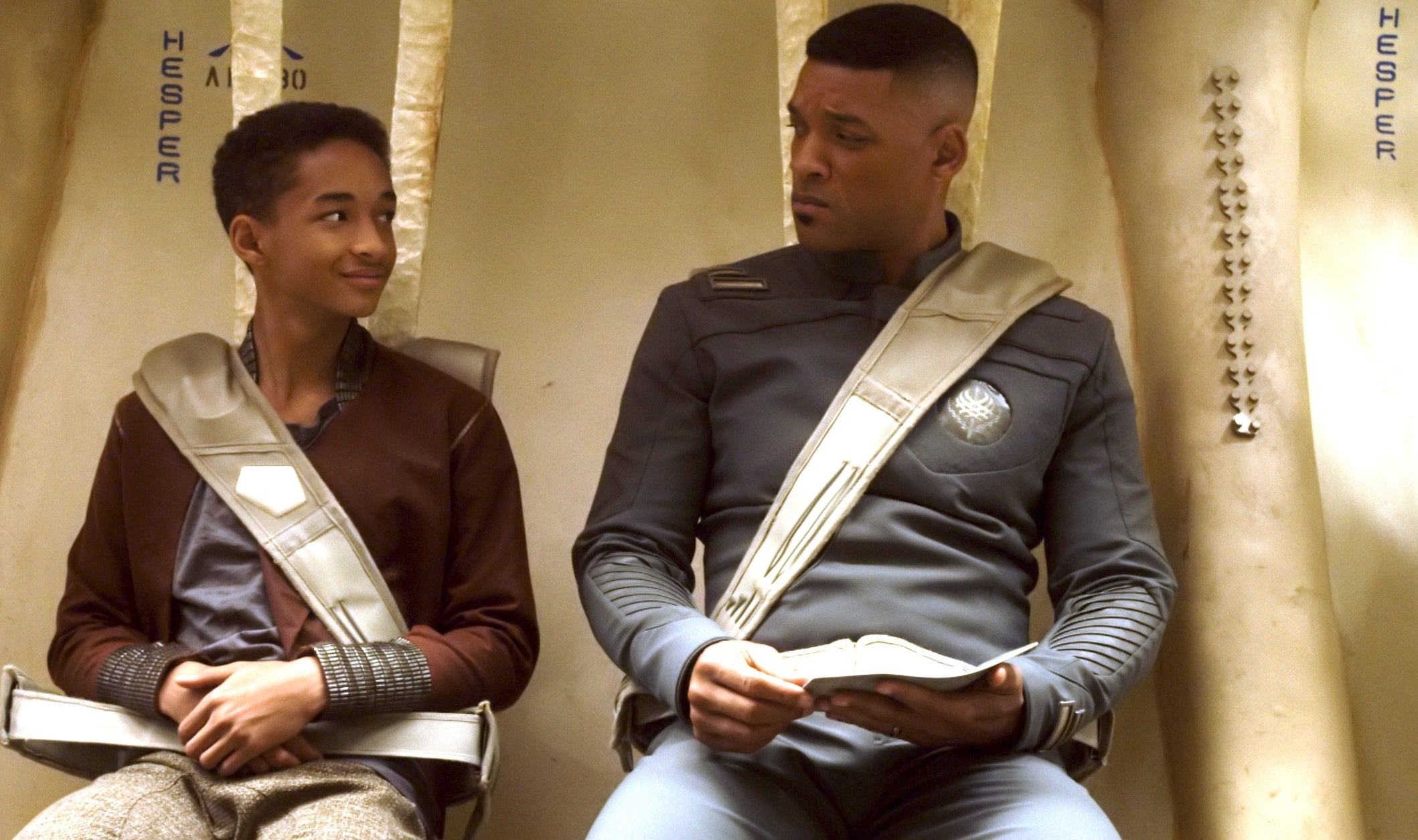 Jaden Smith and Will Smith in futuristic uniforms with seatbelts sit inside a spacecraft and look at each other