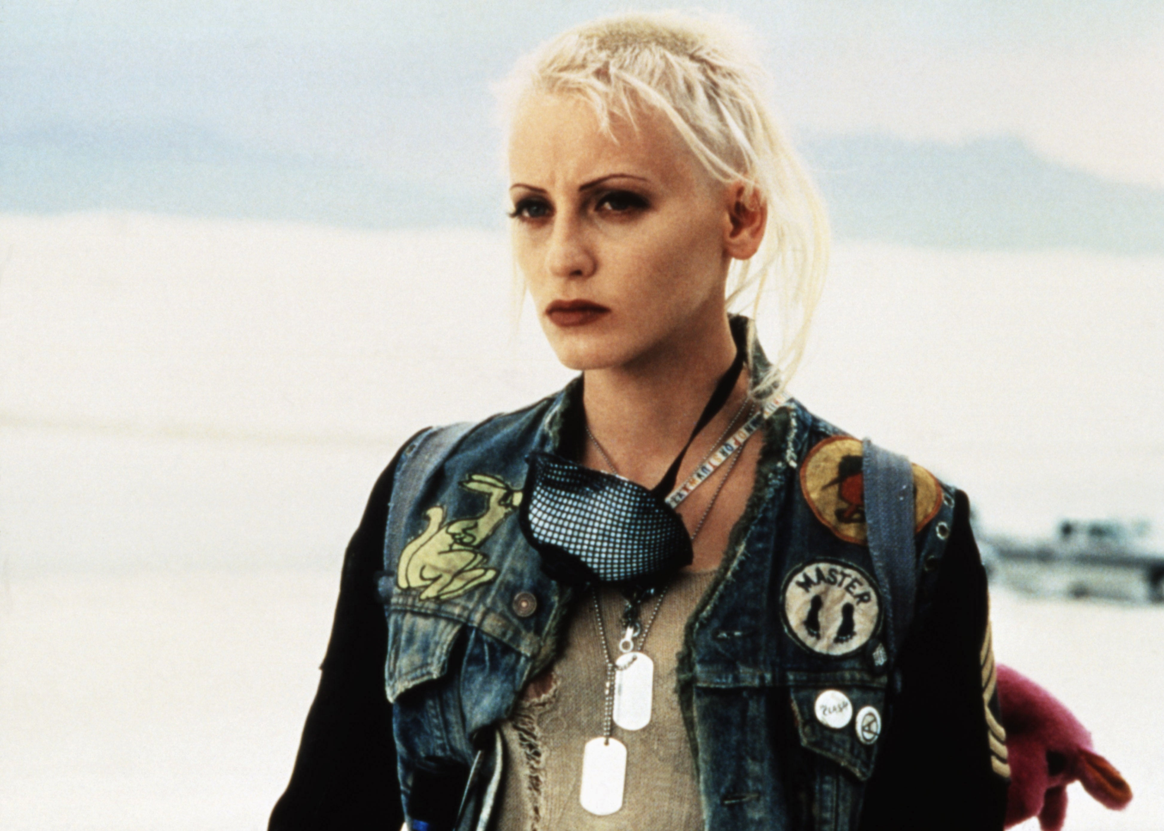 Lori Petty with a punk style, wearing a patched denim vest with pins and dog tags. She has short, light hair and is standing outdoors